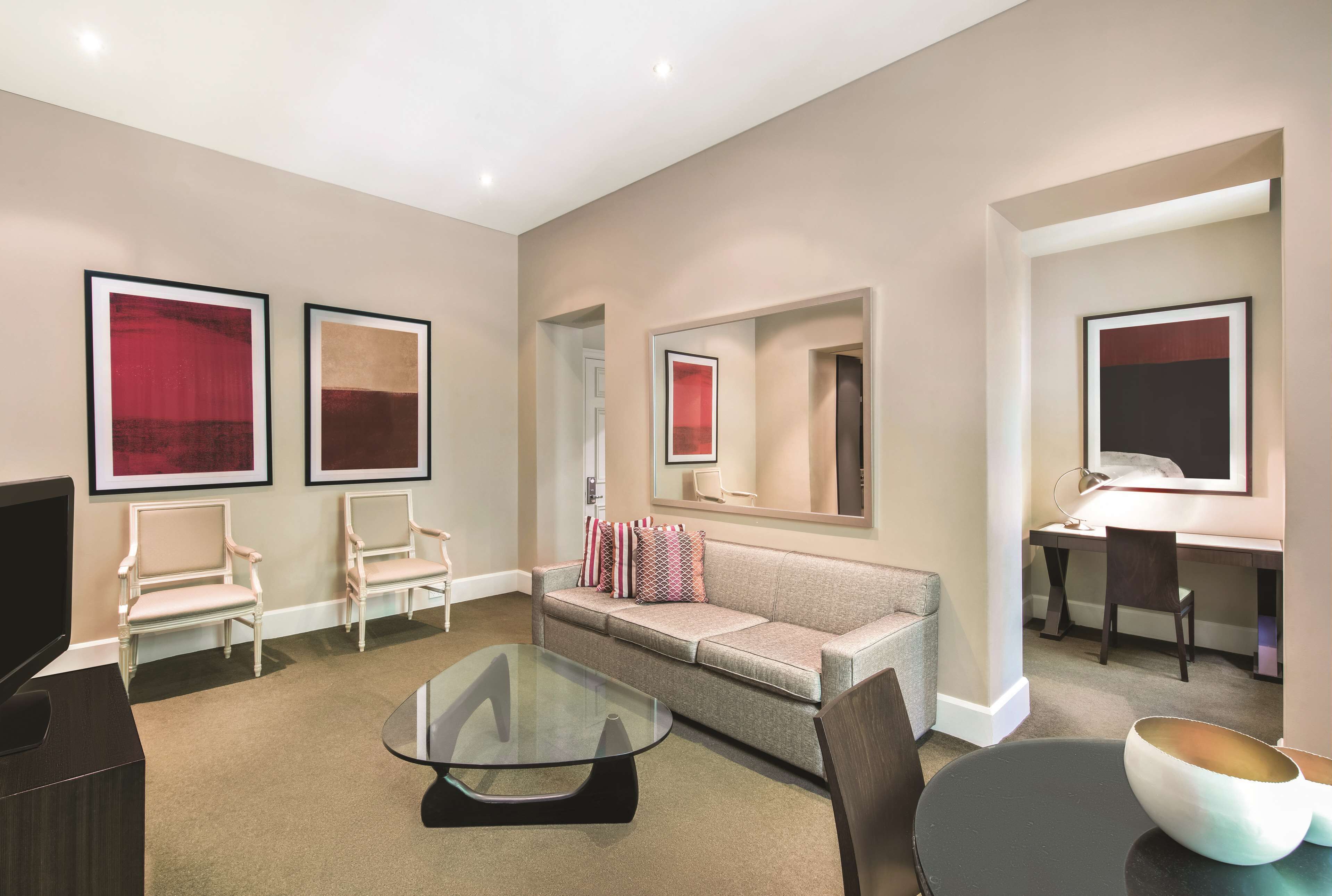 A modern hotel room with a sofa, two armchairs, a glass coffee table, a desk with a chair, and a TV.