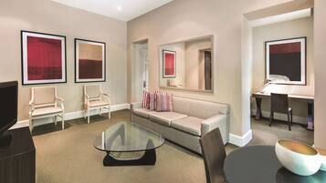 A modern hotel room with a sofa, two armchairs, a glass coffee table, a desk with a chair, and a TV.