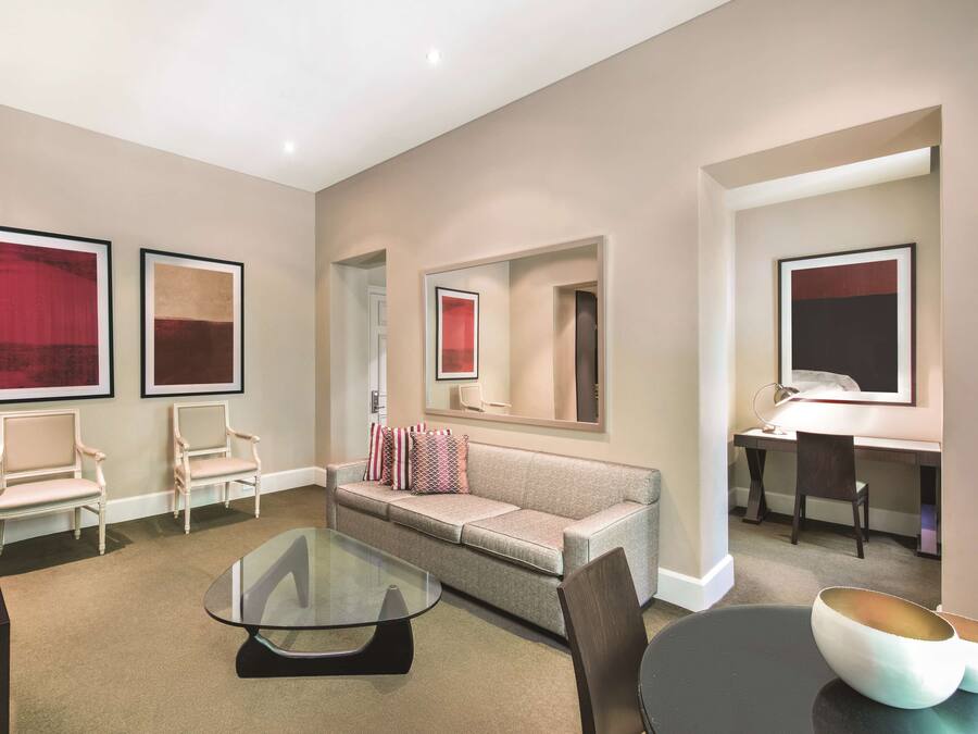 A modern hotel room with a sofa, two armchairs, a glass coffee table, a desk with a chair, and a TV.