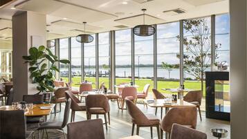 Un vaste espace intérieur aménagé en salle à manger, doté de grandes fenêtres offrant une vue sur un plan d’eau et la verdure environnante. Des tables sont disposées avec des chaises, et une grande plante est visible au premier plan.