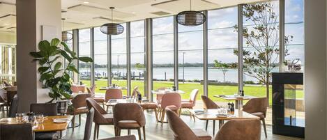 A spacious indoor dining area with large windows offering a view of a body of water and greenery. Tables are set with chairs, and a large plant is visible in the foreground.