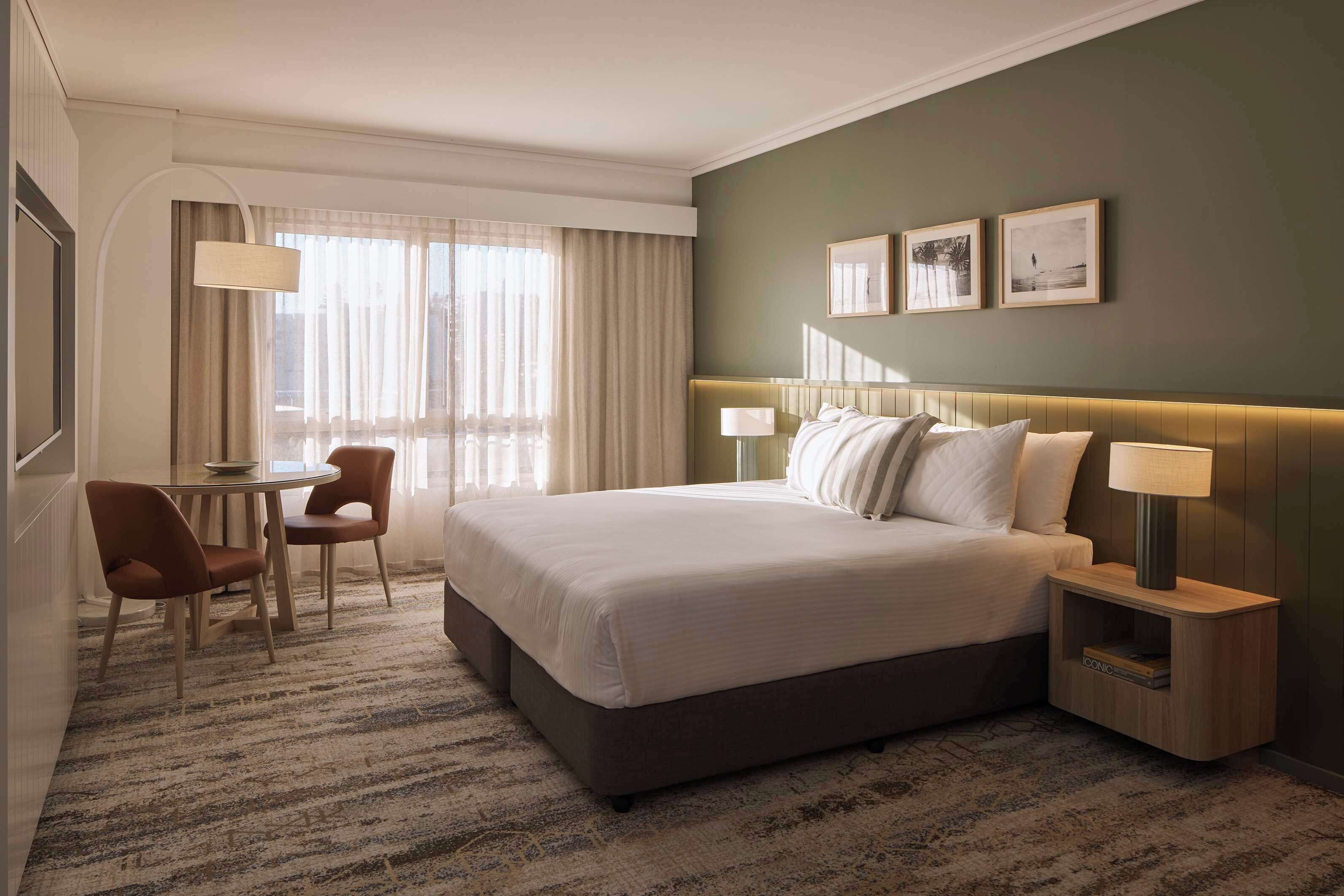 Habitación de hotel con una cama grande, una mesita de noche con lámpara, una pequeña mesa con sillas y vistas a la ciudad desde la ventana.
