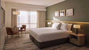 Habitación de hotel con una cama grande, una mesita de noche con lámpara, una pequeña mesa con sillas y vistas a la ciudad desde la ventana.
