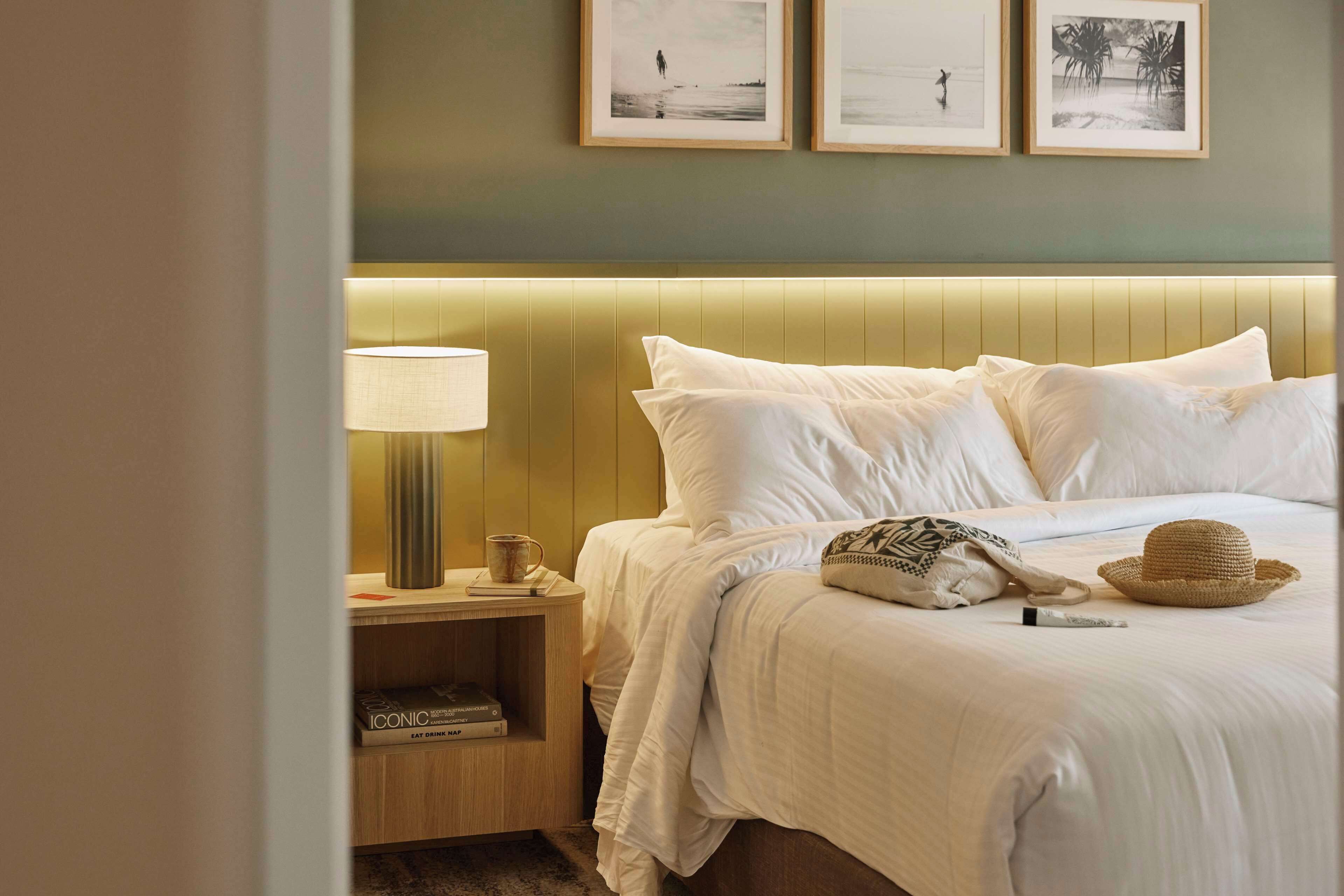 A neatly made bed with white linens, a lamp, and a hat on the side.