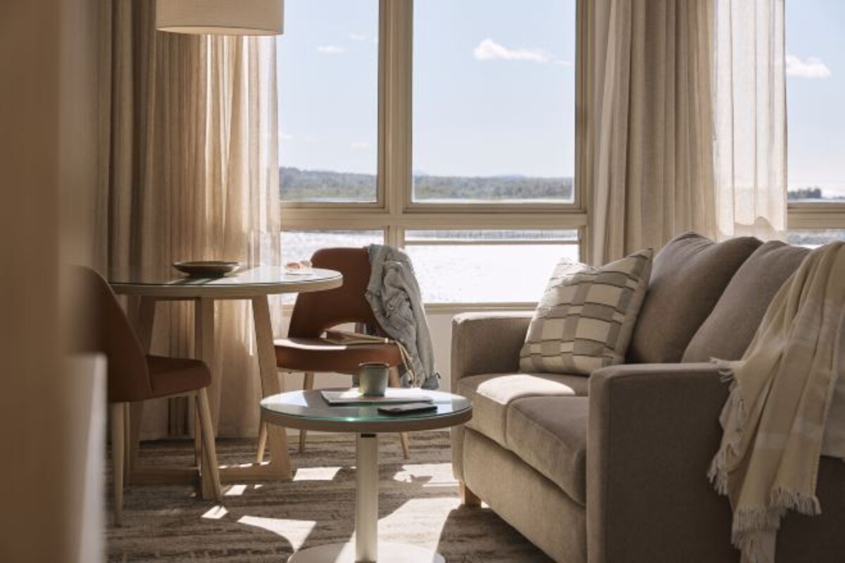 A living room with a sofa, a small round table, a chair, and a view of a body of water through large windows.