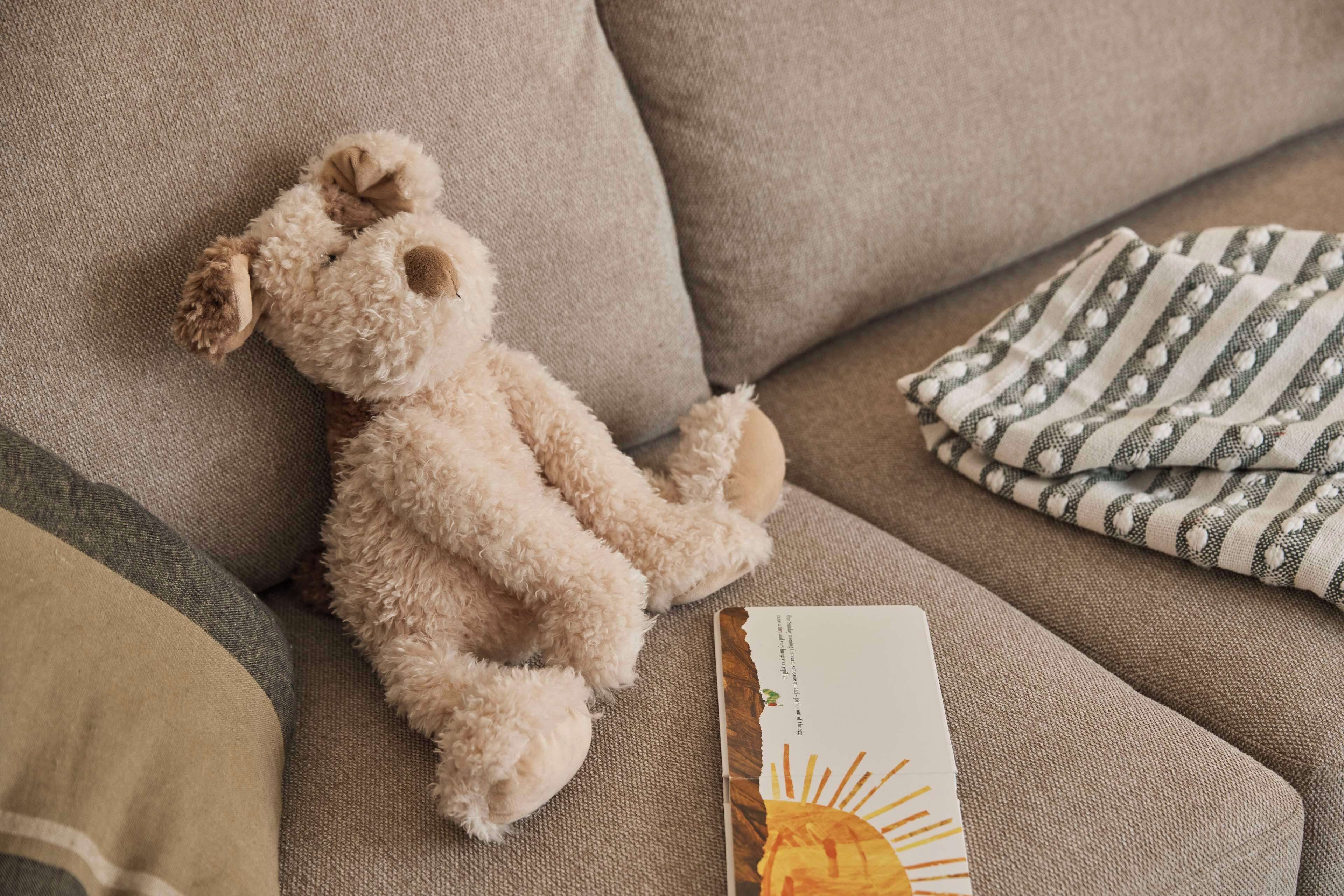 Un sofá color beige con un osito de peluche, un paño a rayas y un libro.
