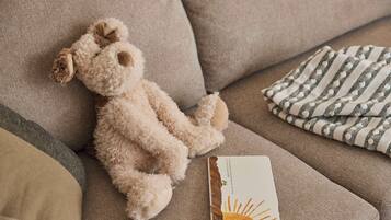 Un sofá color beige con un osito de peluche, un paño a rayas y un libro.