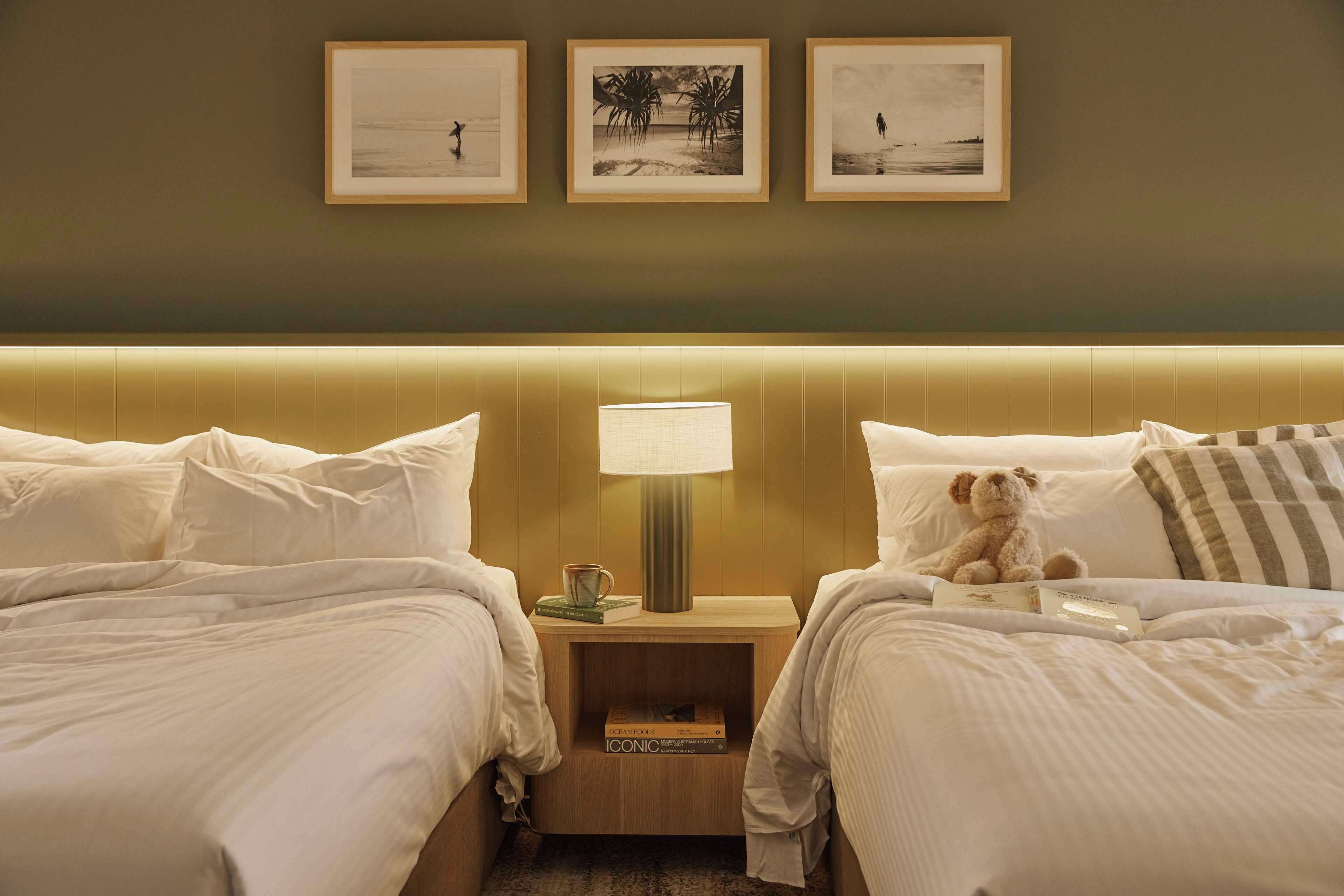 Habitación de hotel con dos camas, una mesita de noche con lámpara y tres cuadros en la pared.