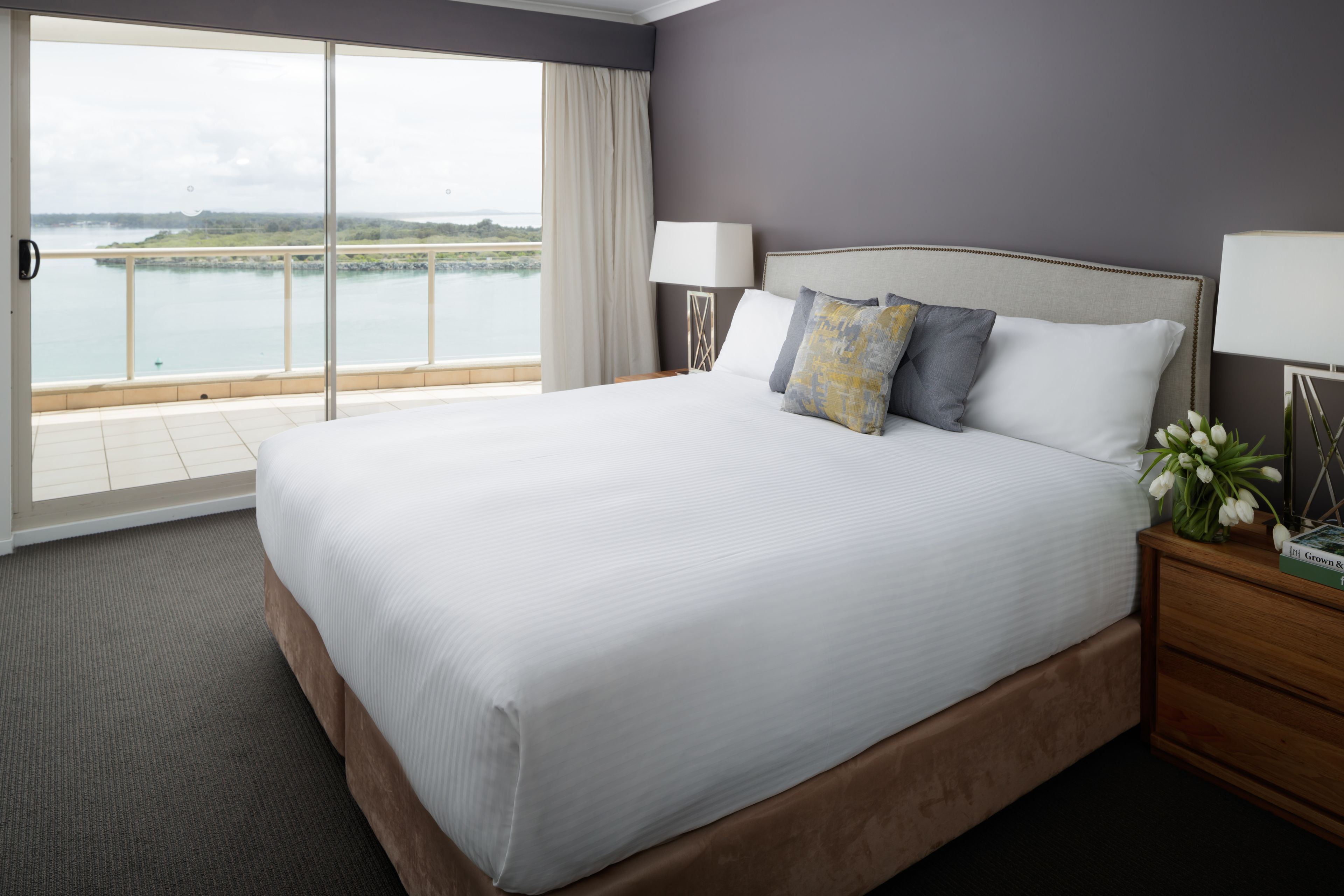 Una cama grande con sábanas blancas, una mesita de noche de madera con una lámpara, y vista a un cuerpo de agua a través de una puerta corrediza.