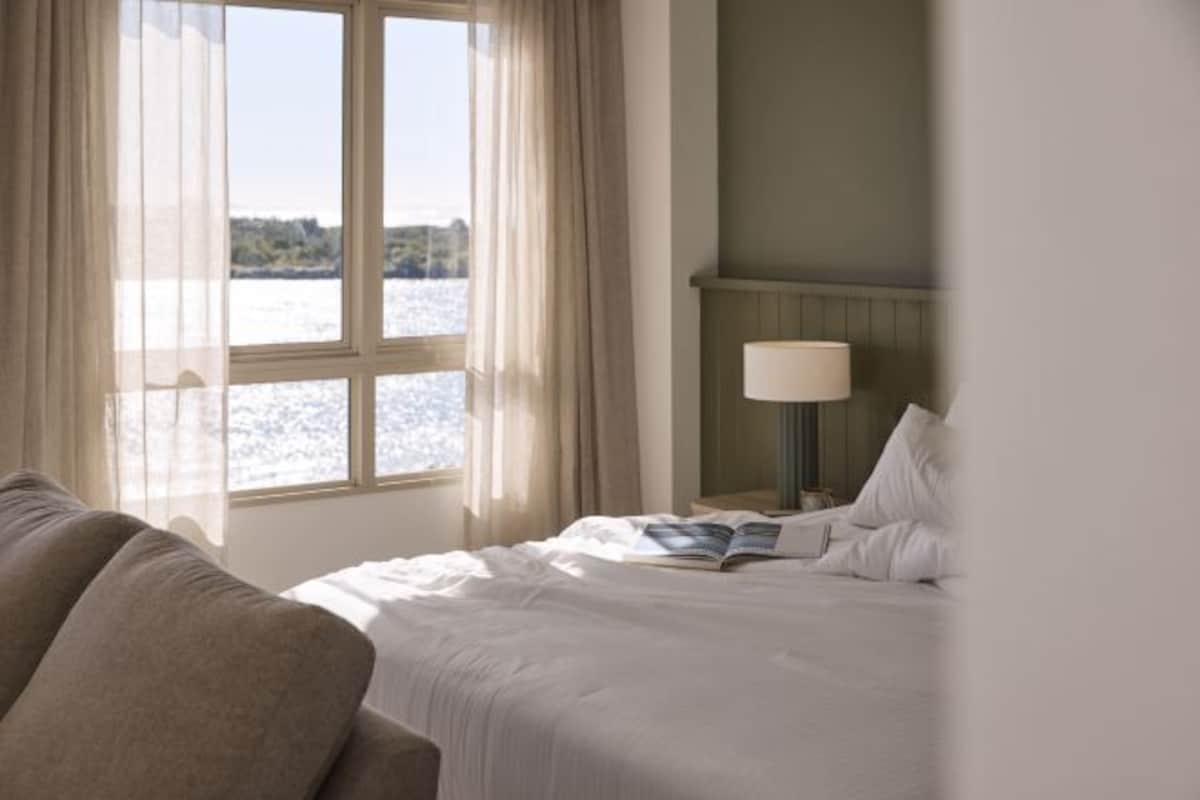 A hotel room with a bed, a nightstand, a lamp, and a view of a body of water through a window.