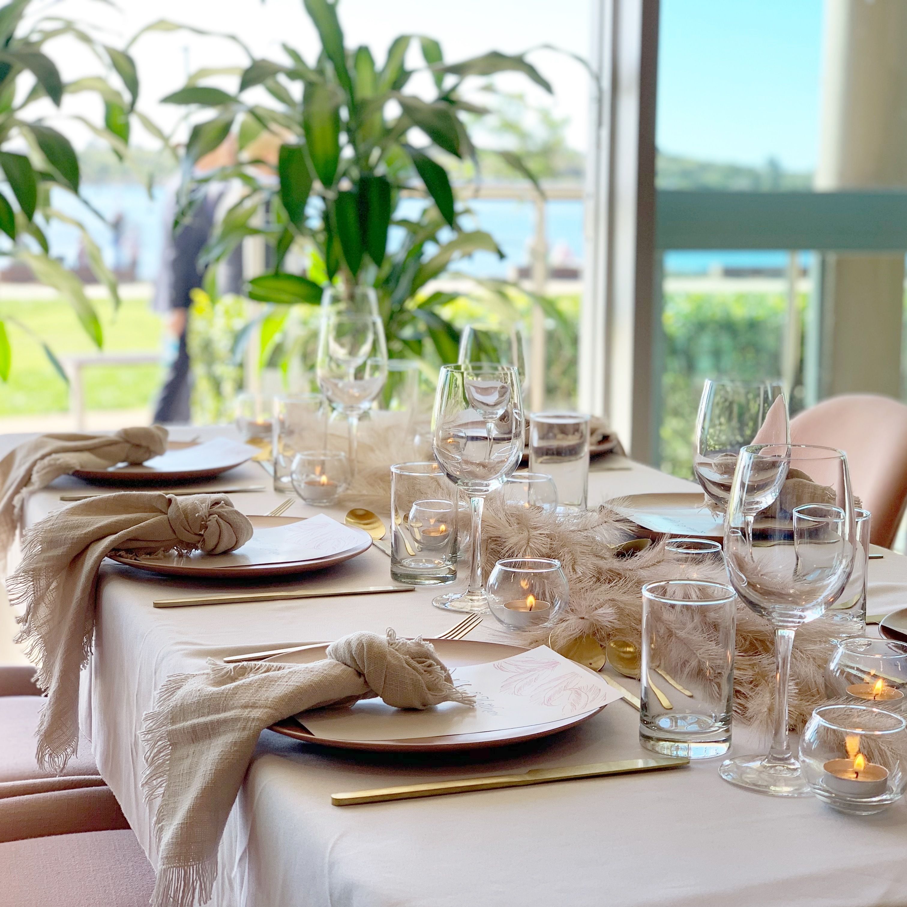 A table set for dining with clear glassware, candles, and a view of greenery outside.