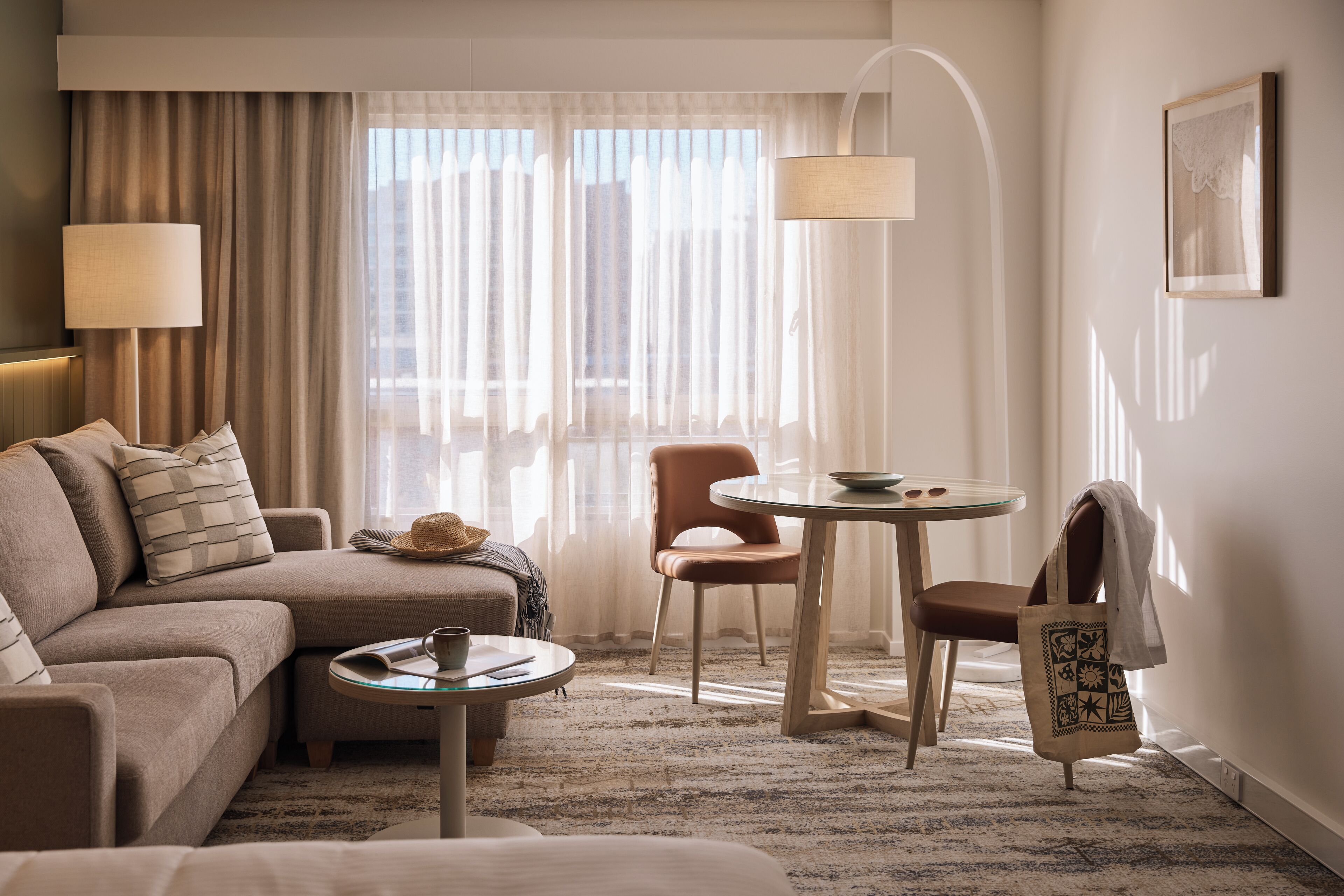 A hotel room with a sofa, a small round table, a chair, a lamp, and a window with curtains.