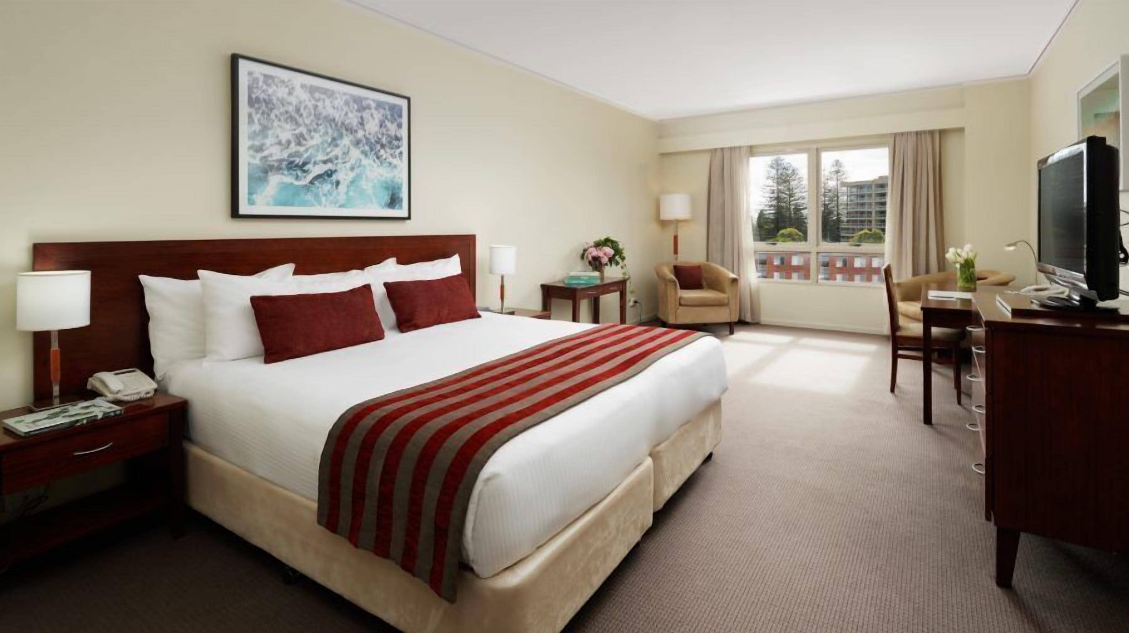 A hotel room with a large bed, a desk with a TV, a chair, a desk lamp, and a window with a view of buildings.