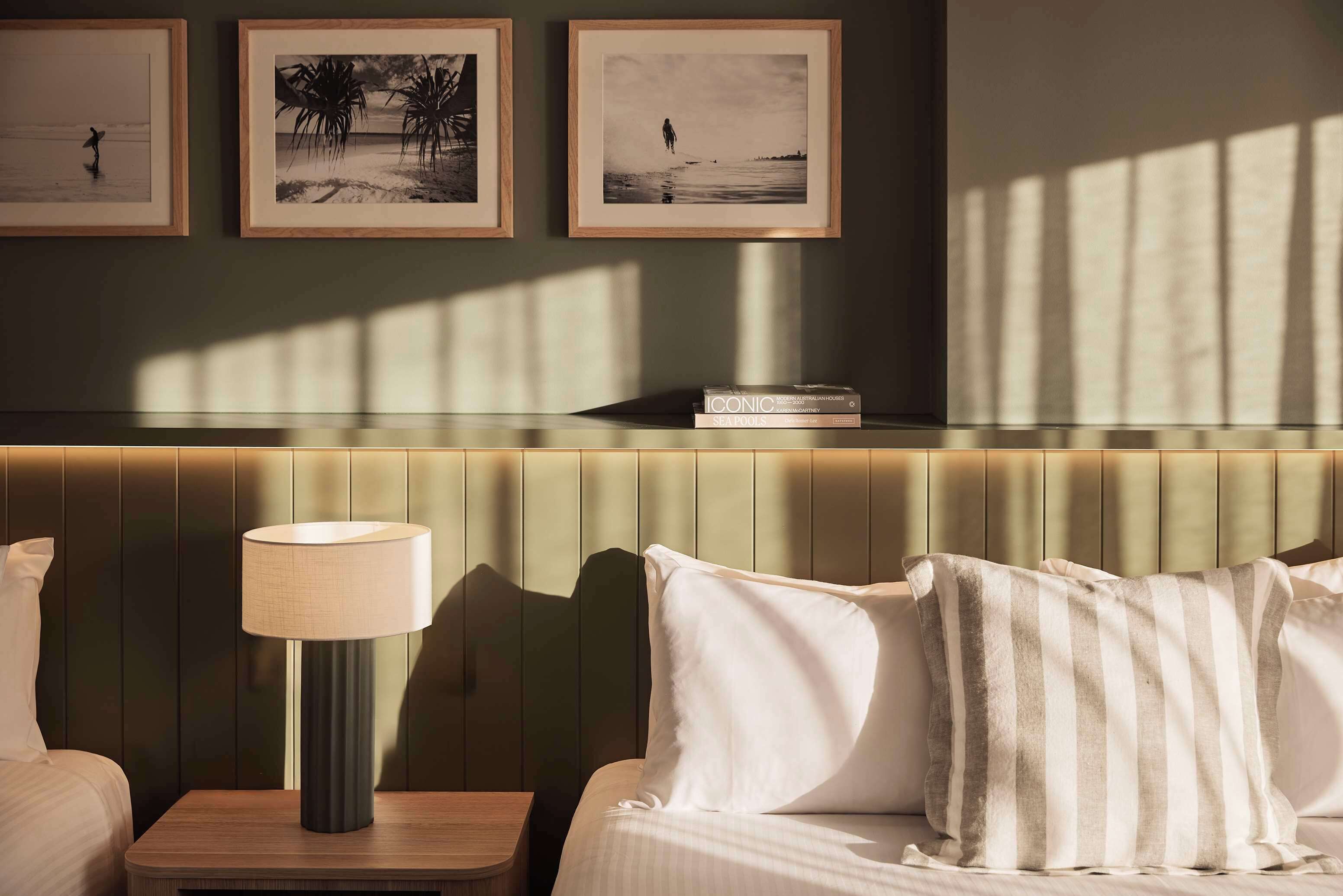 A hotel room with a bed, a bedside table with a lamp, a bookshelf, and framed pictures on the wall.