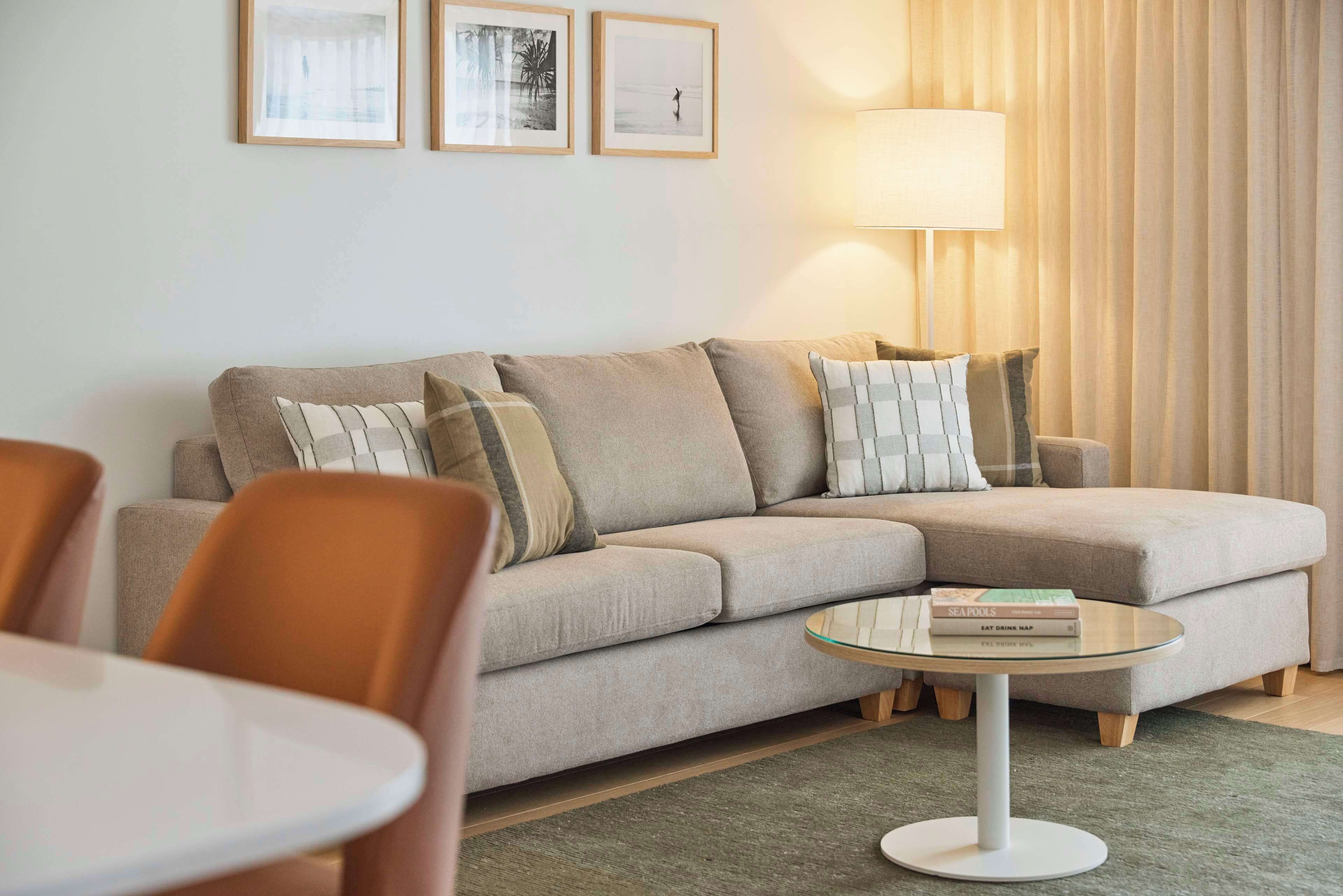 A living room with a beige sofa, a glass coffee table, and a lamp. There are framed pictures on the wall and a window with curtains.