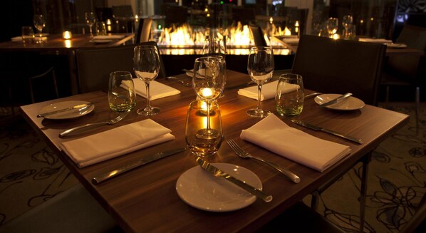 Mesa de comedor preparada para cuatro personas, con copas de vino, platos y cubiertos, y un candelabro encendido como centro de mesa.