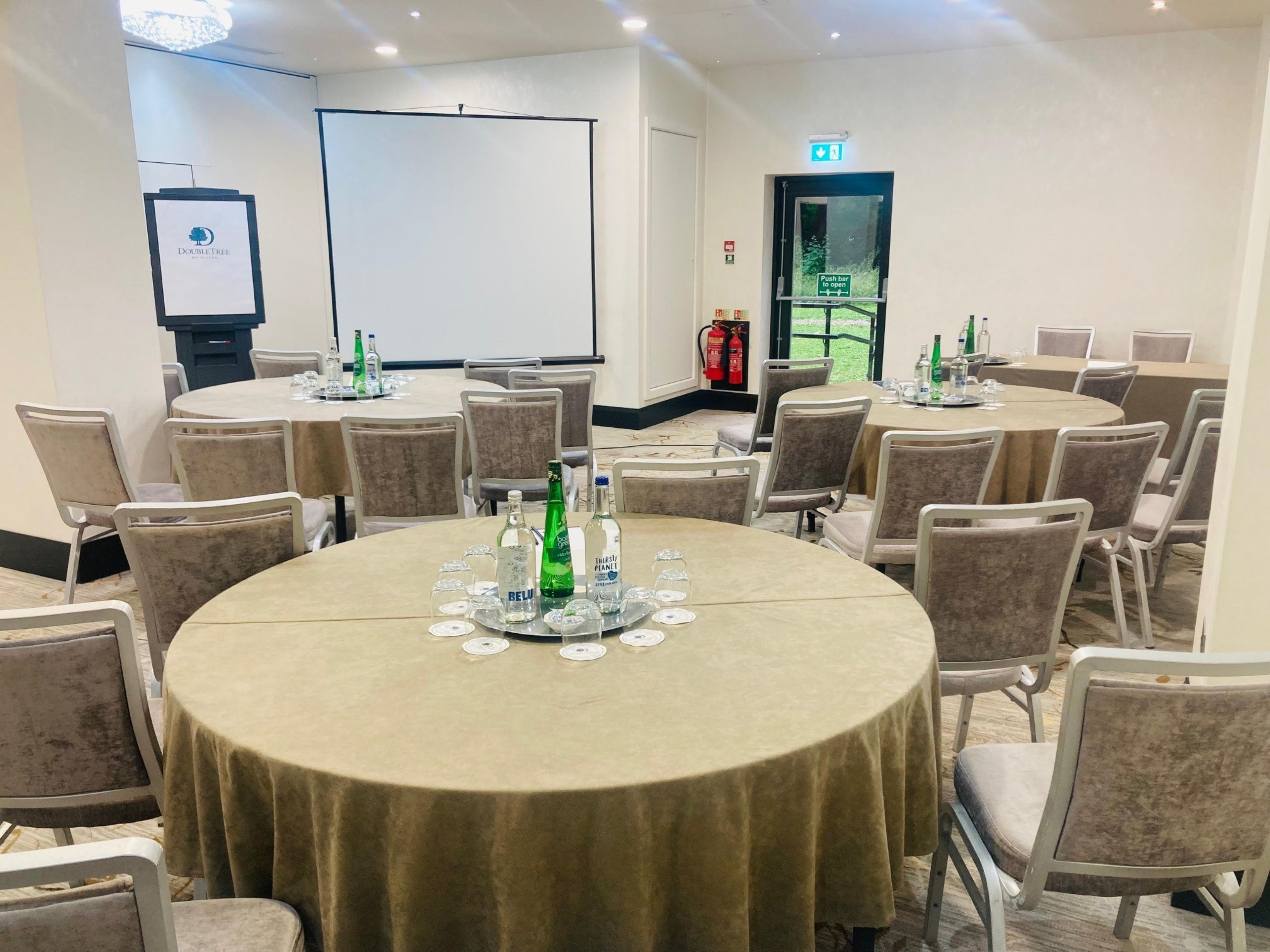 Un salón de conferencias con mesas redondas, sillas y una pantalla para proyector.