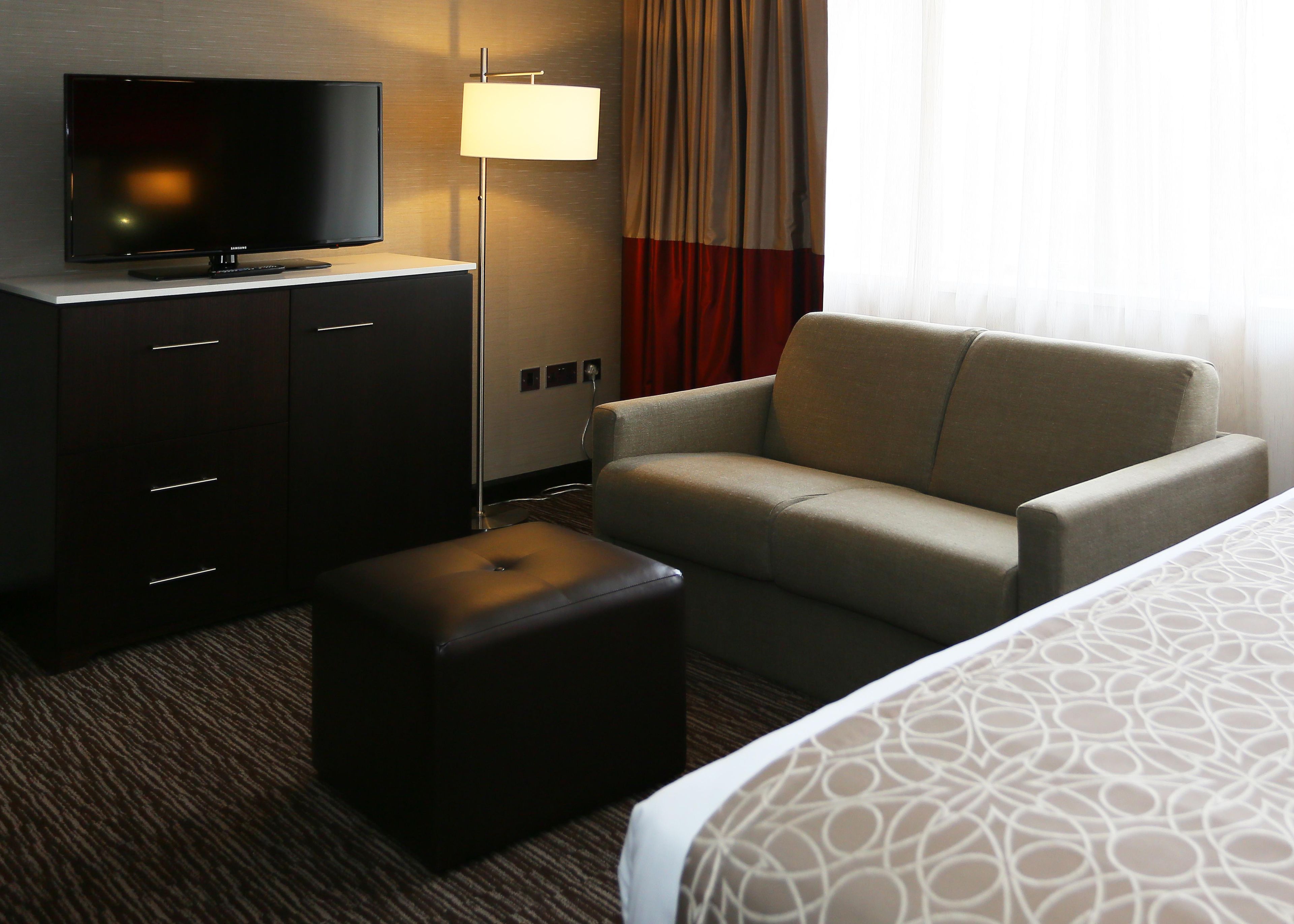 A hotel room with a flat-screen TV on a cabinet, a sofa, an ottoman, and a bed with patterned bedding.