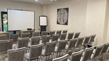 A conference room with rows of chairs, a projector screen, a whiteboard, and a decorative wall piece.