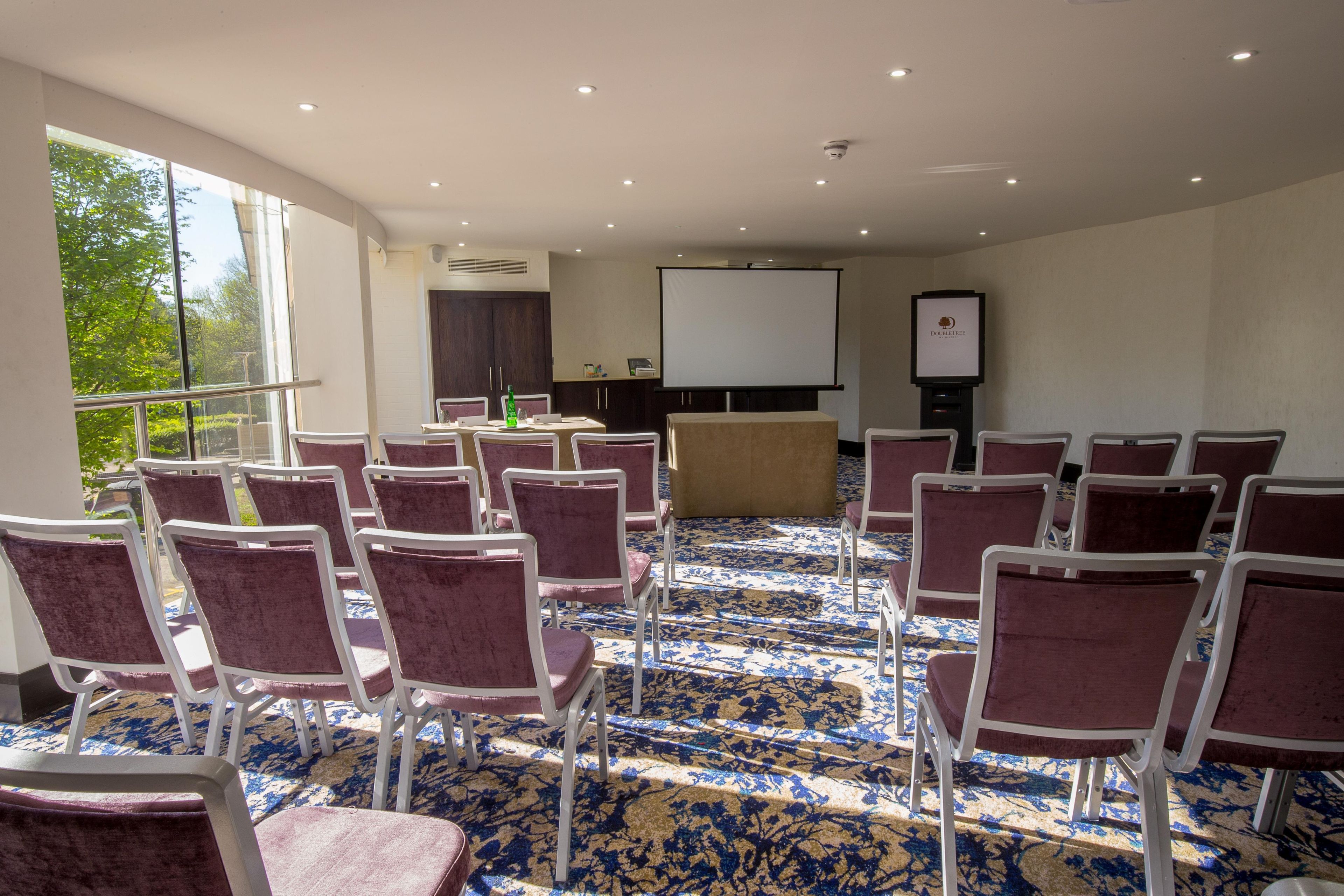 Un salón de conferencias con filas de sillas, una pantalla de proyector y una zona de presentaciones.