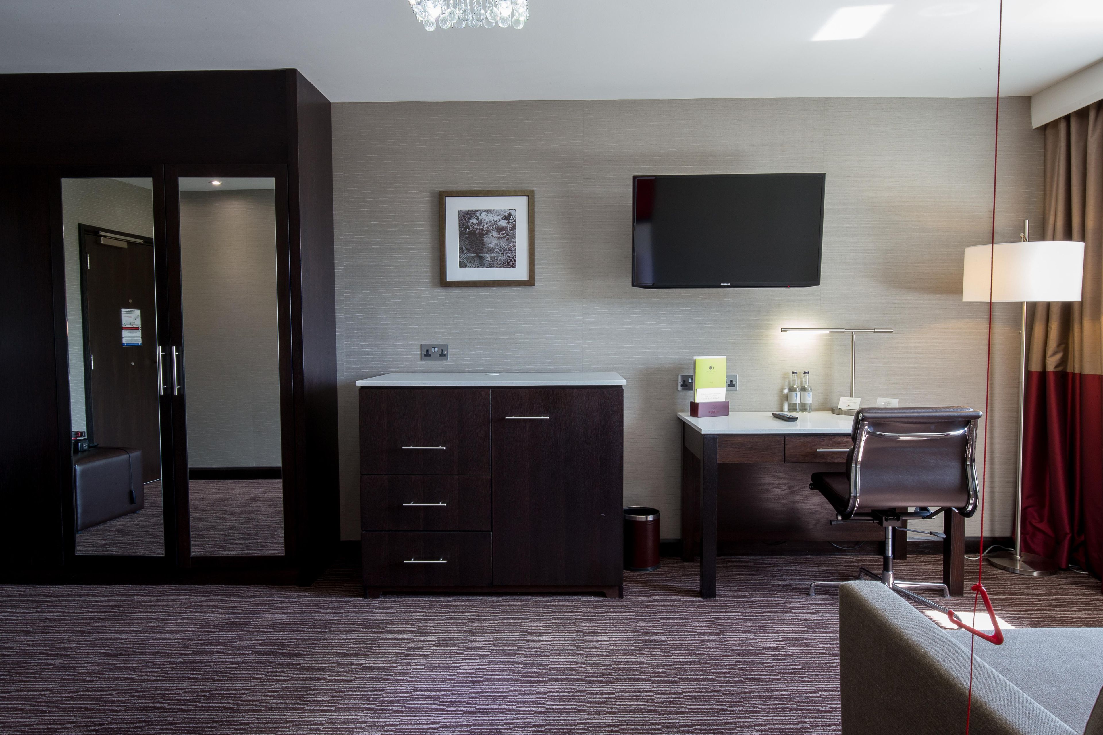 A hotel room with a desk, a flat-screen TV, a large mirror, a wardrobe, a chair, and a lamp.