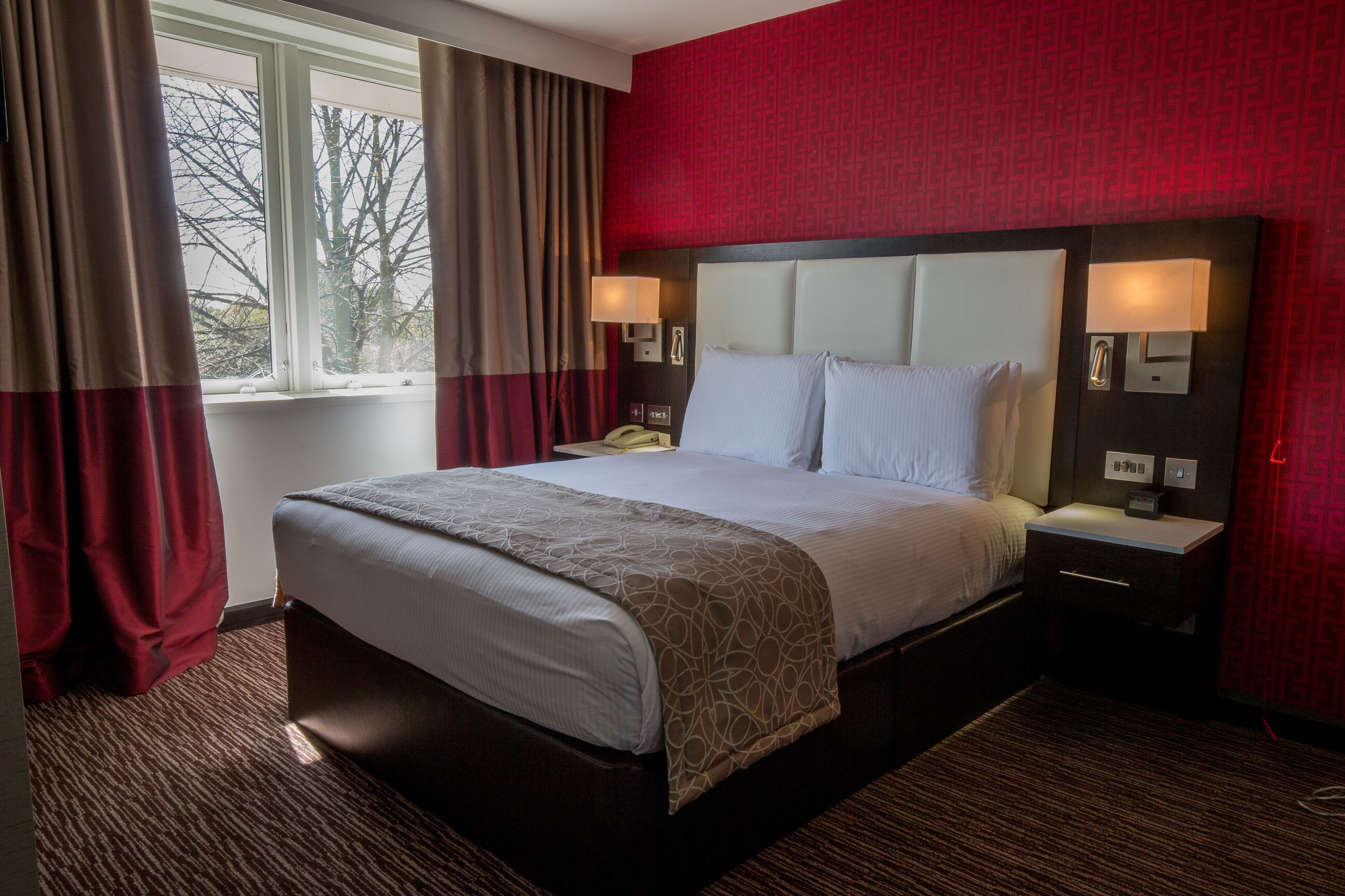 Ein Hotelzimmer mit einem großen Bett, zwei Nachttischen, einem Fenster mit Vorhängen und einer roten Wand mit Muster.