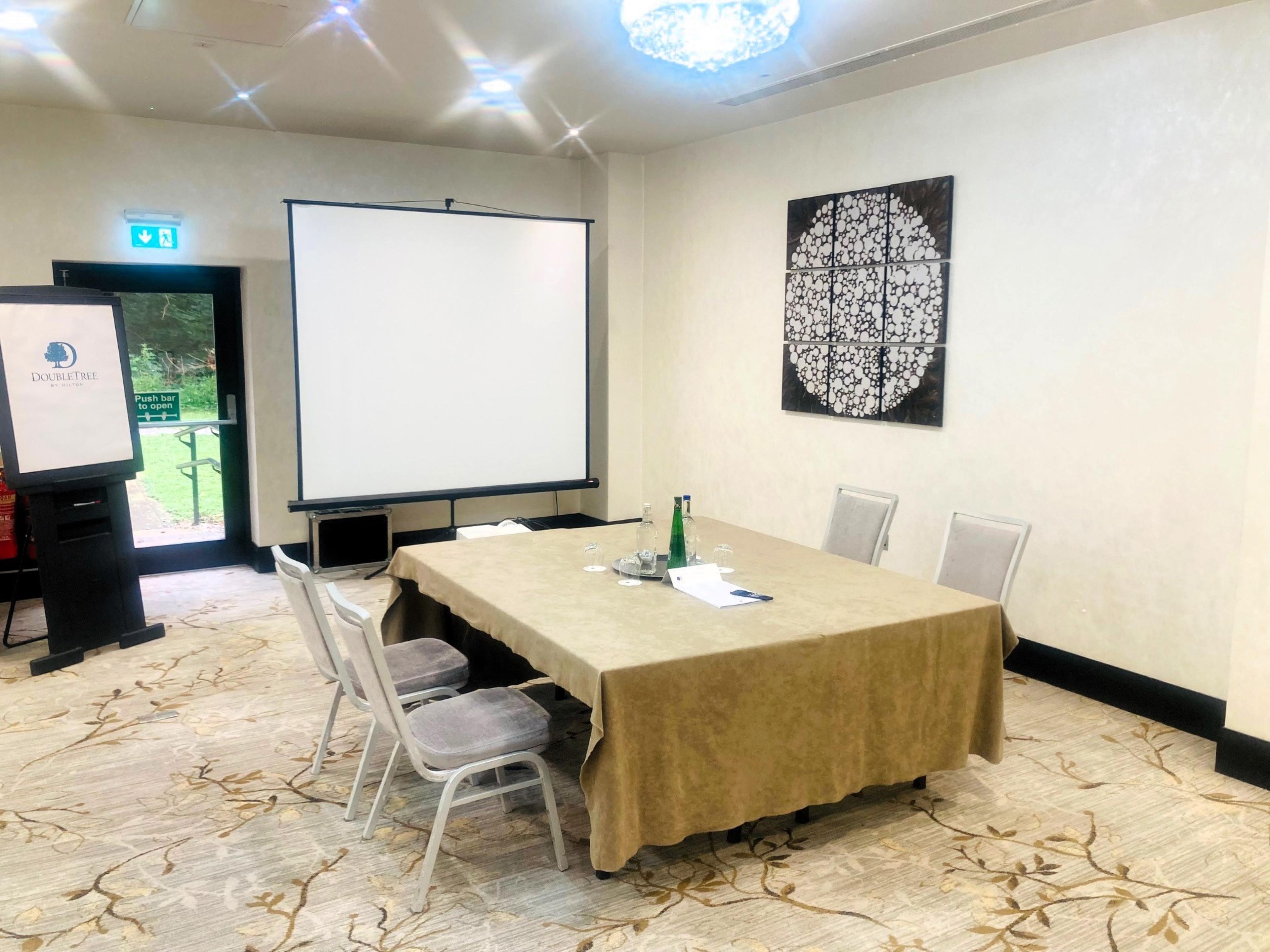 Un salón de conferencias con una larga mesa, sillas, una pantalla de proyector, una barra de empuje y un adorno en la pared.