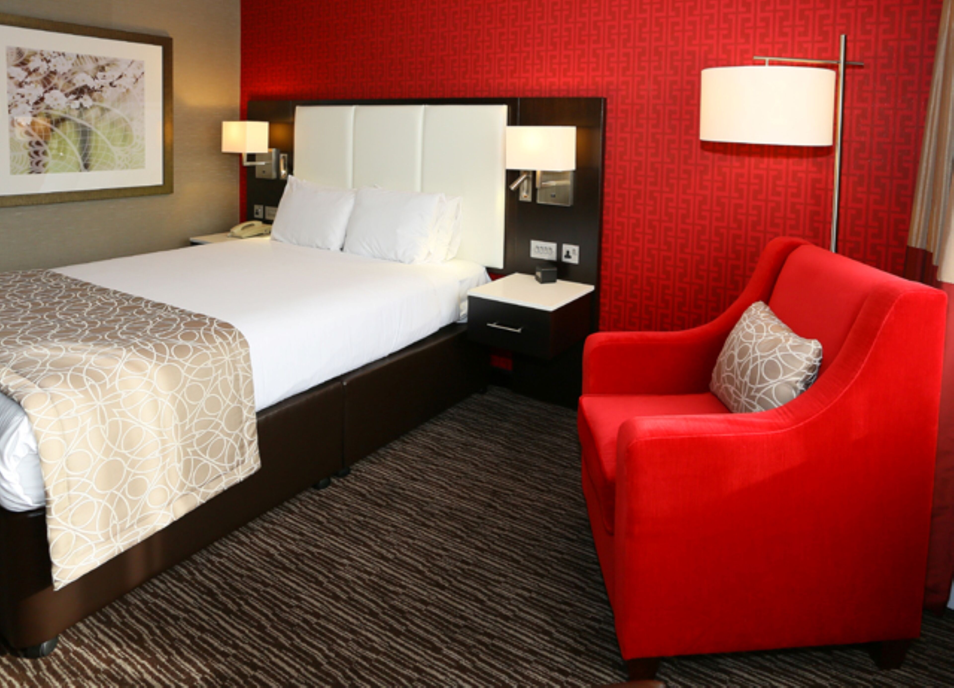 A hotel room with a large bed, a nightstand, a red armchair, and a wall-mounted lamp.