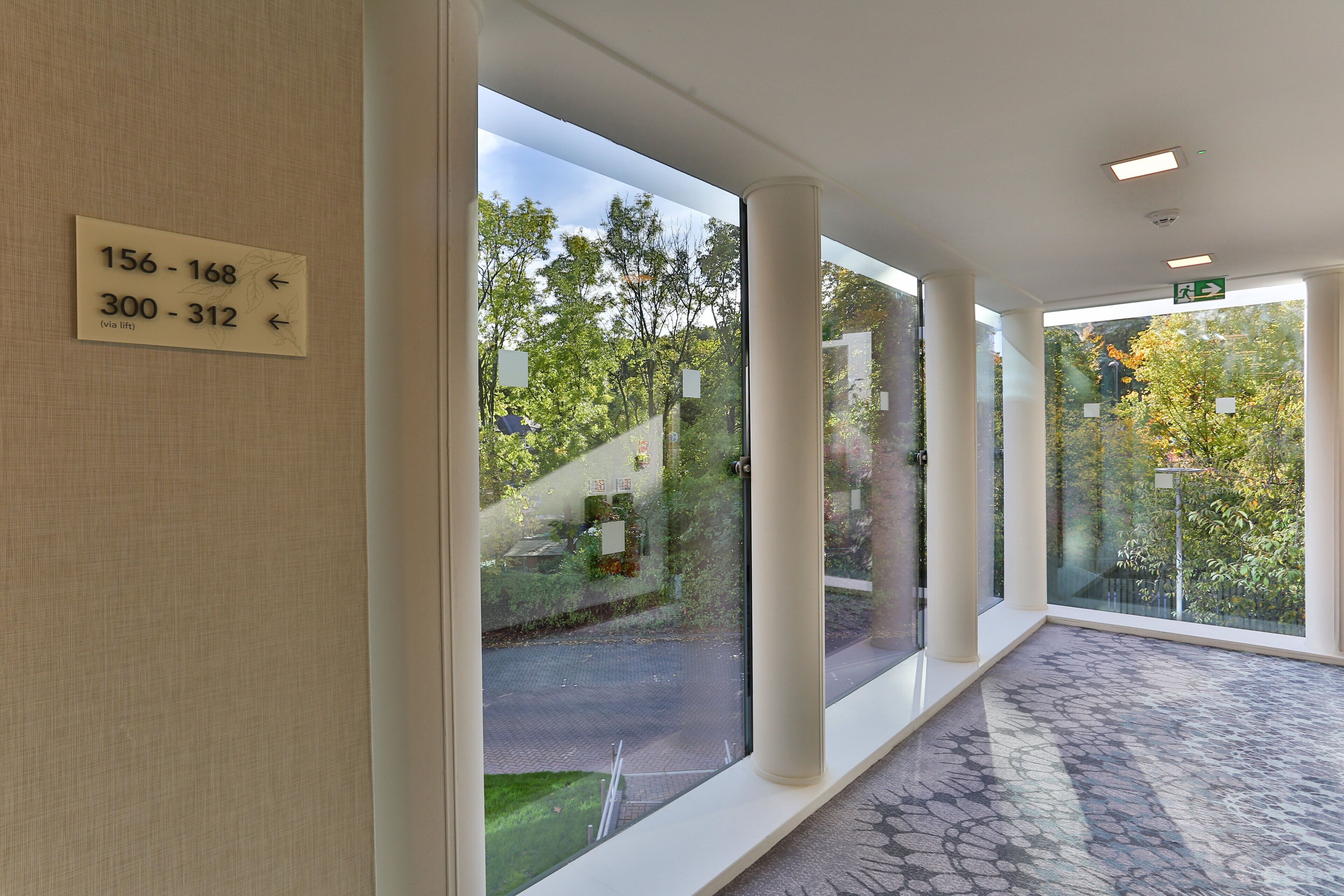 A corridor with large windows, a sign indicating room numbers, and a patterned floor.