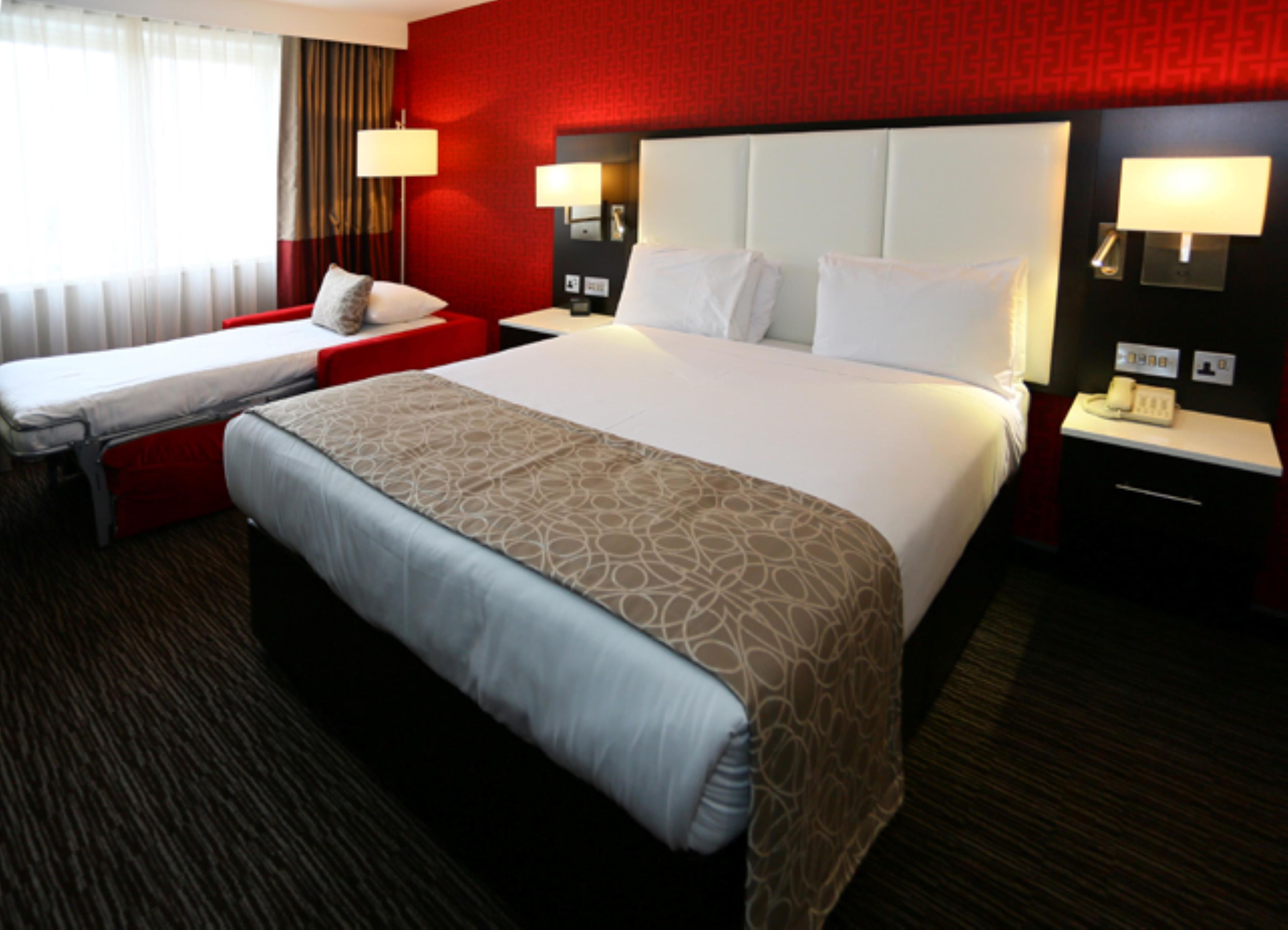 A hotel room with a large bed, a smaller bed, two bedside tables with lamps, and a red accent wall.