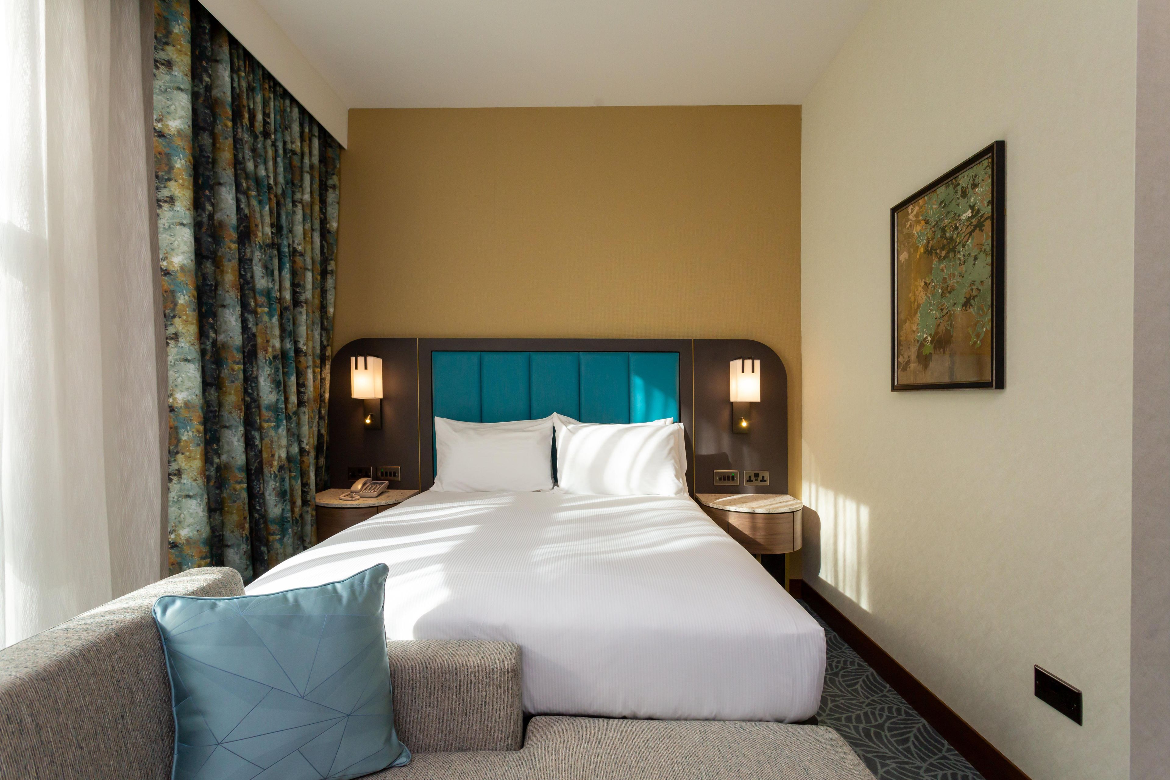 Una habitación de hotel con cama, dos mesitas de noche, un sofá, una cortina estampada y un cuadro en la pared.