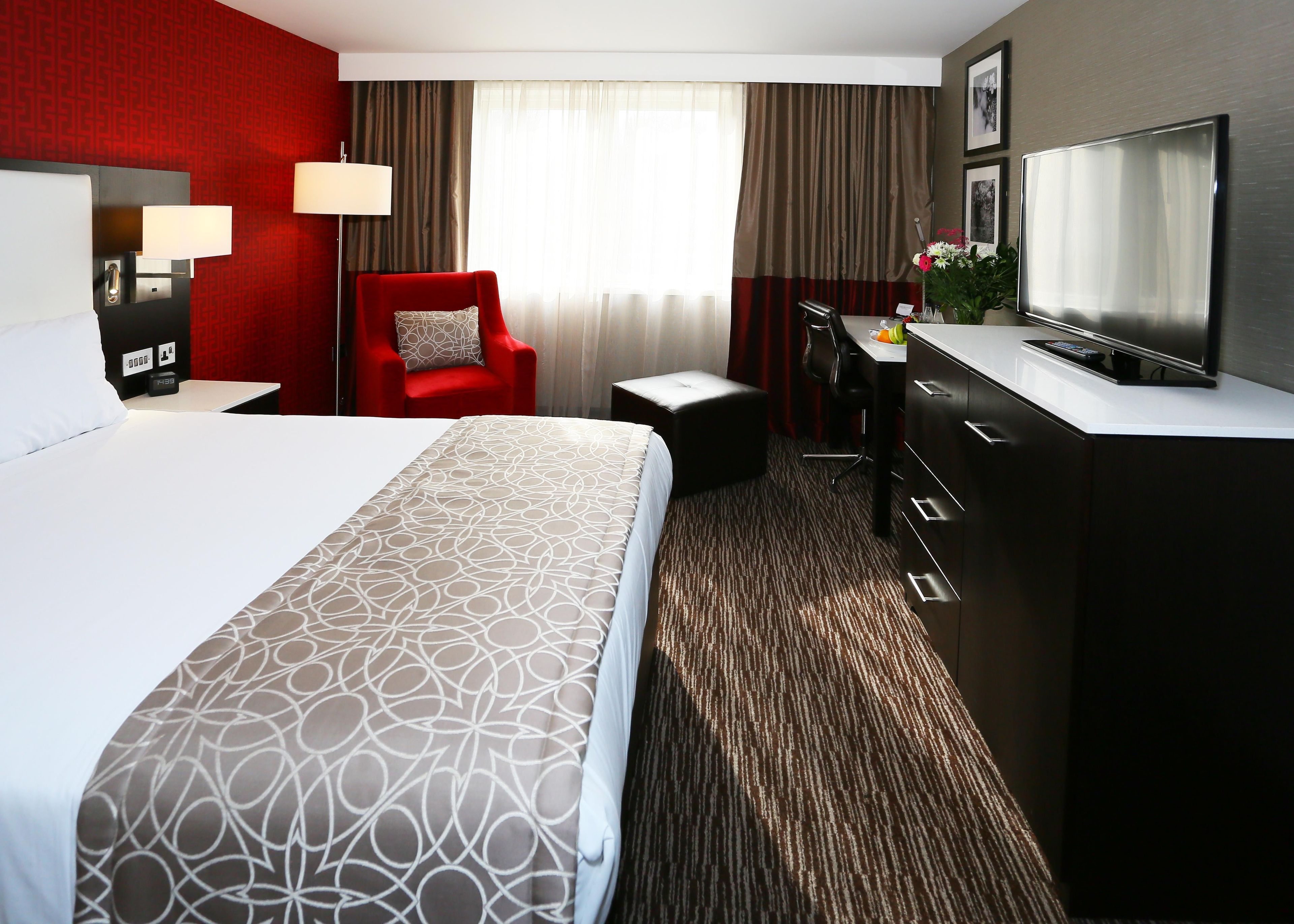 A hotel room with a large bed, a desk with a chair, a television, and a red armchair.
