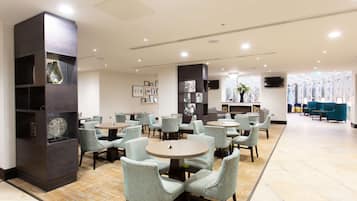 Un hall d’hôtel moderne, avec des tables et des chaises rondes, une grande structure en bois sombre dotée d’étagères, et un téléviseur fixé au mur.