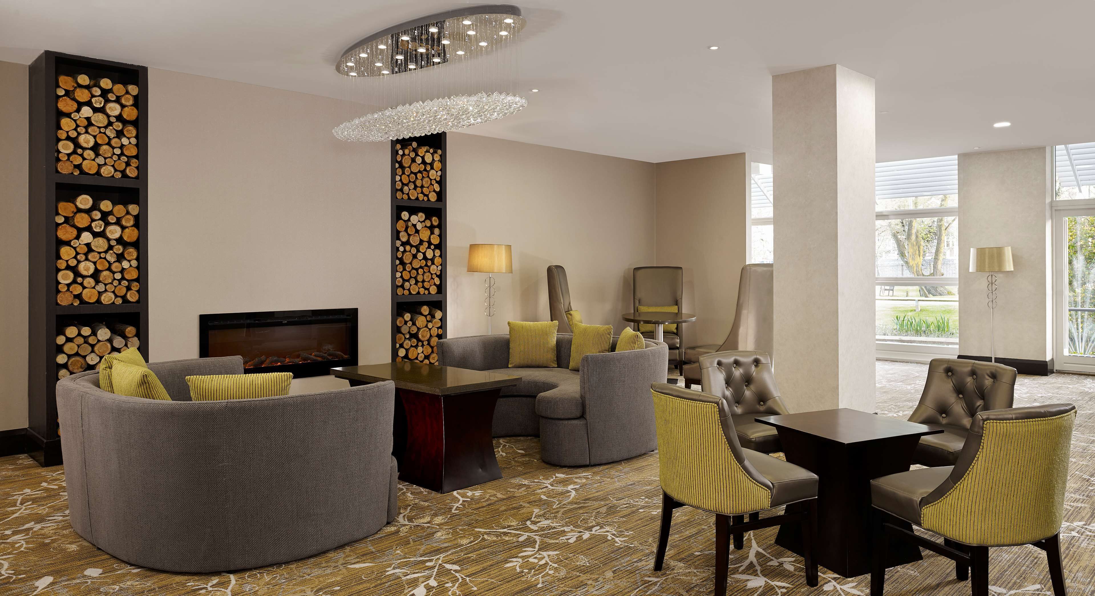 Um lobby moderno de hotel com um sofá curvo, uma mesa de jantar com cadeiras, uma lareira e uma parede decorativa com toras.