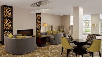 A modern hotel lobby with a curved sofa, a dining table with chairs, a fireplace, and a decorative wall with logs.