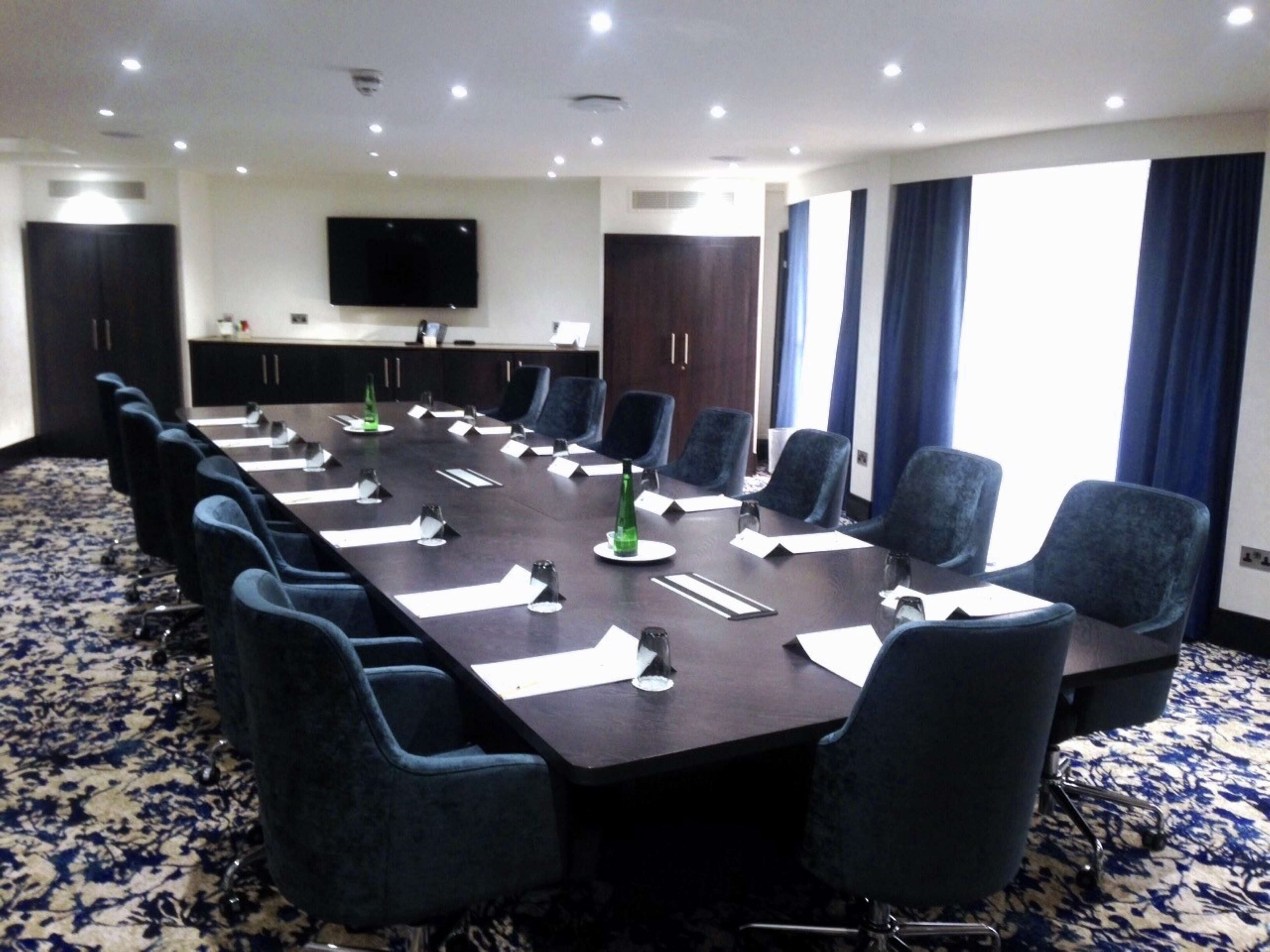 A conference room with a long table, chairs, a flat-screen TV, and a patterned carpet.