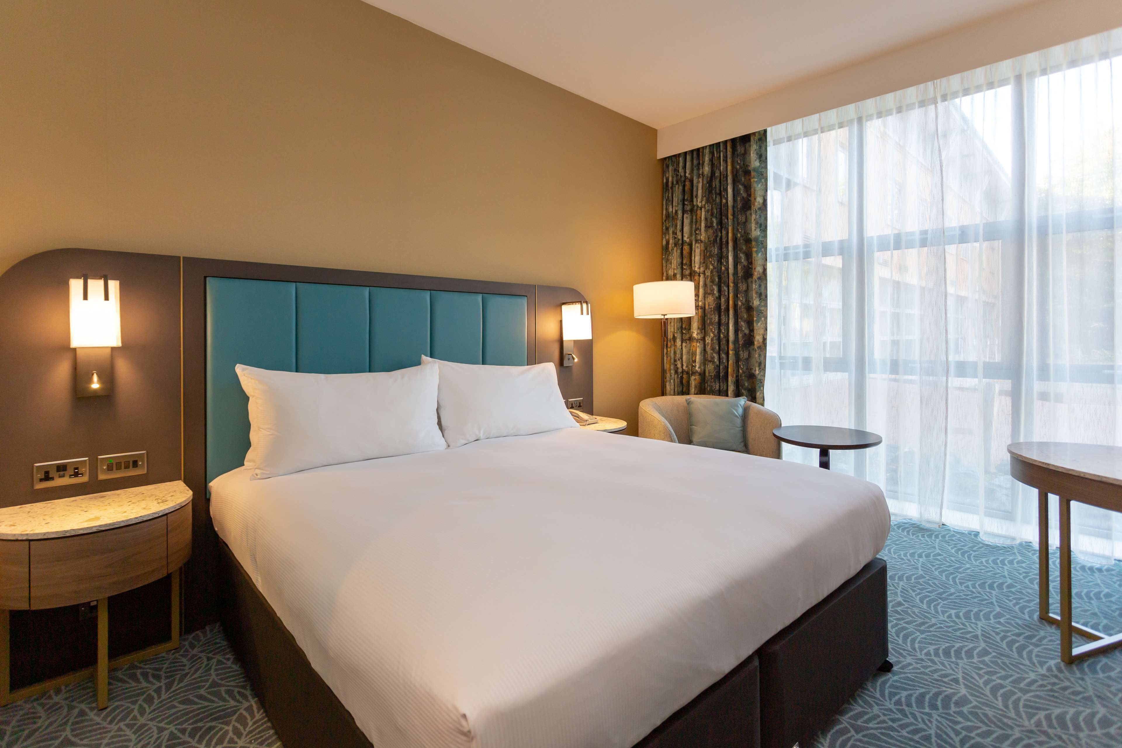 A hotel room with a large bed, two bedside tables, a chair, a small table, and a large window with curtains.