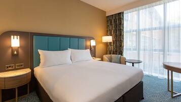 A hotel room with a large bed, two bedside tables, a chair, a small table, and a large window with curtains.