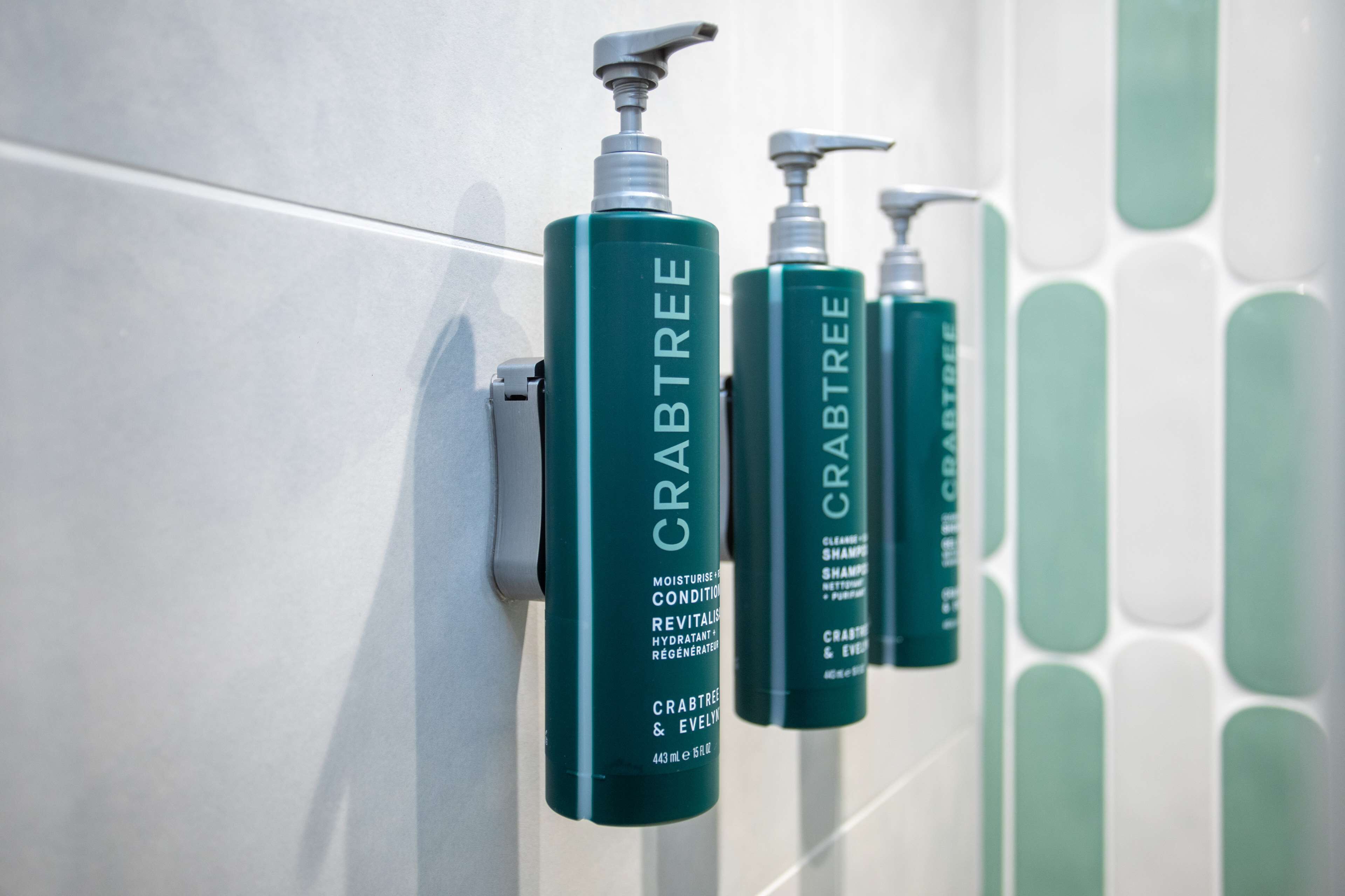 Uma fileira de garrafas verdes com a etiqueta 'CRABTREE' numa parede revestida de azulejos.