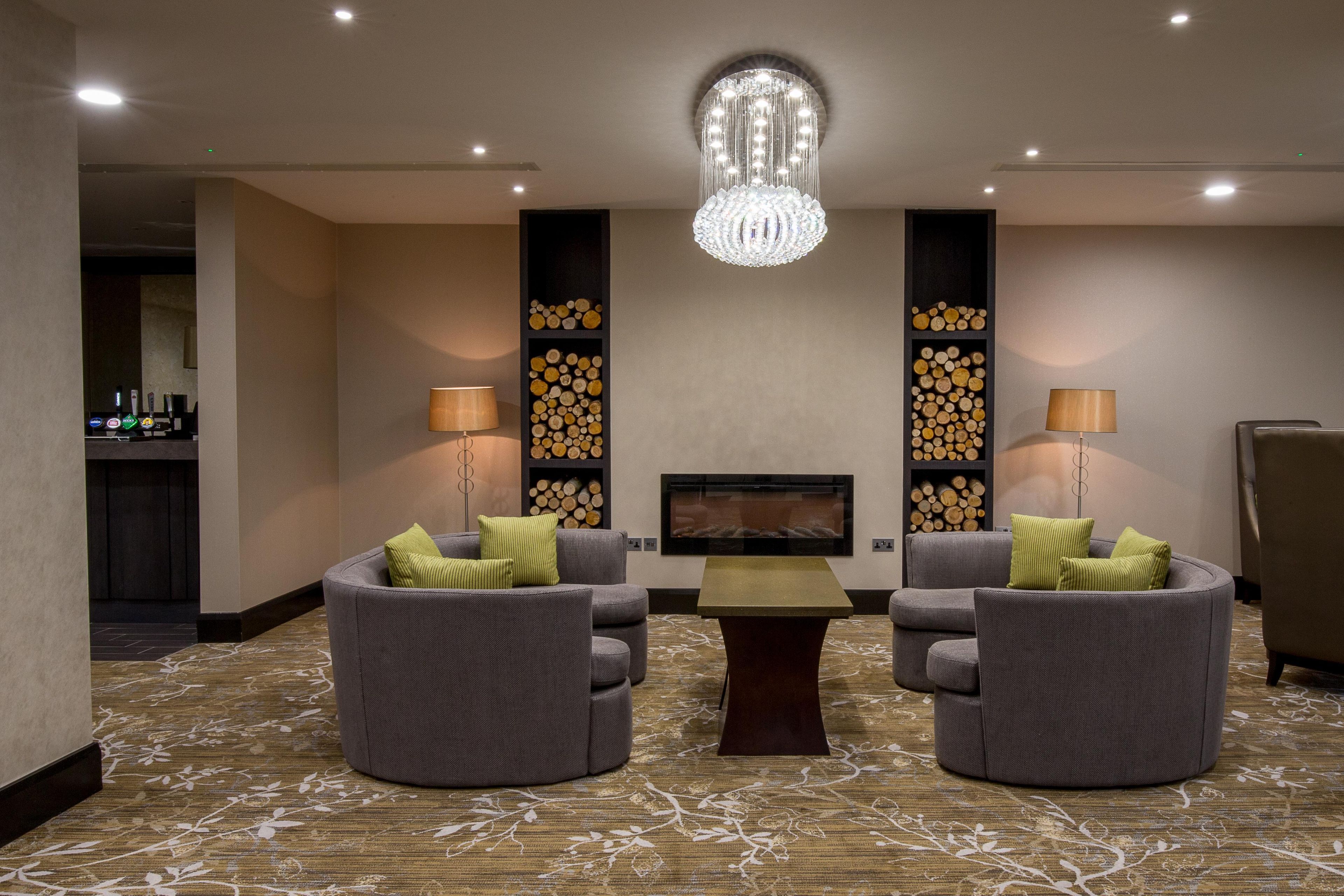 A hotel room with two grey armchairs, a small table, a chandelier, and a fireplace.