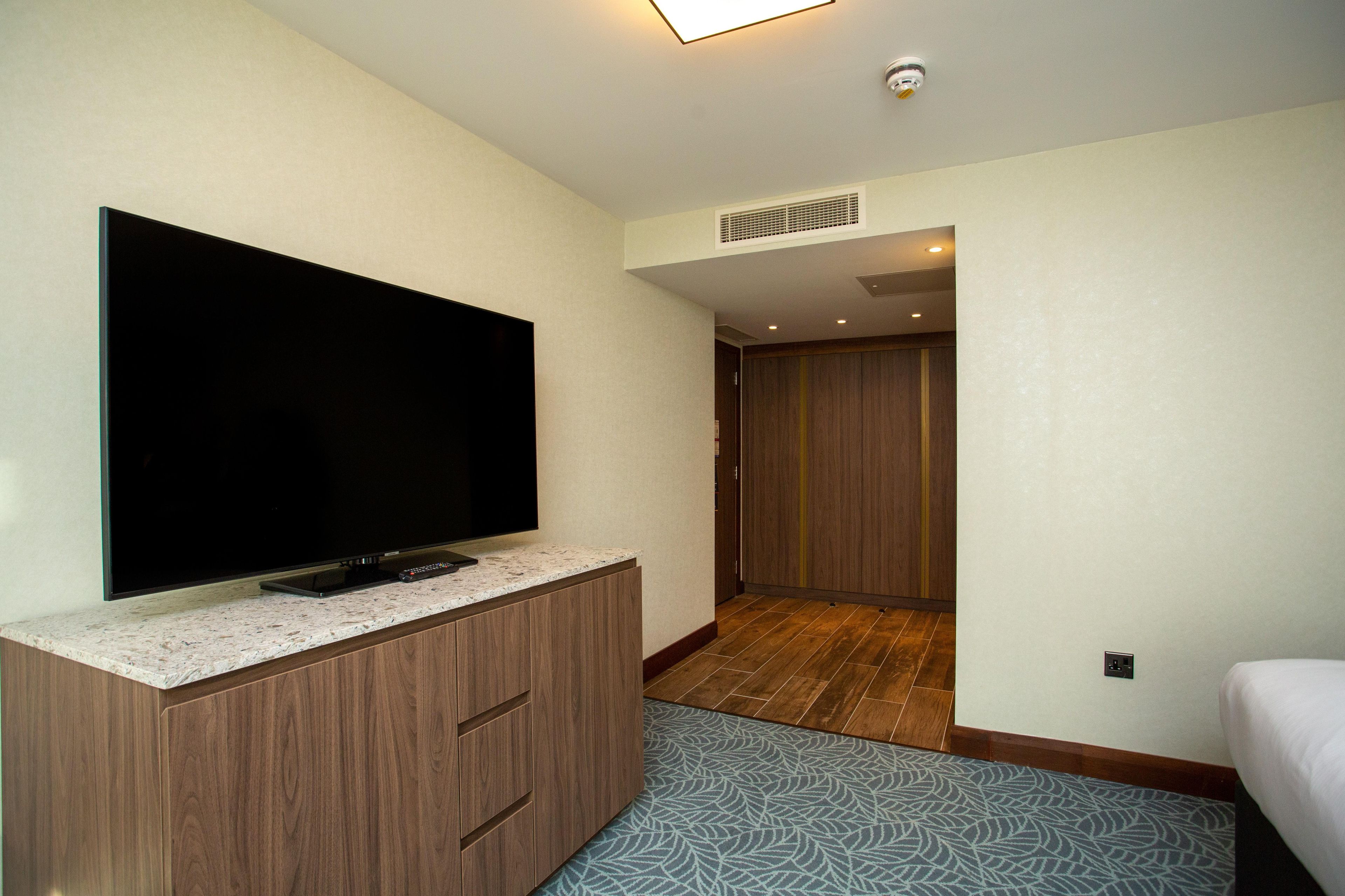 Habitación de hotel con televisor de pantalla plana sobre un mueble de madera, alfombra estampada y una cama.