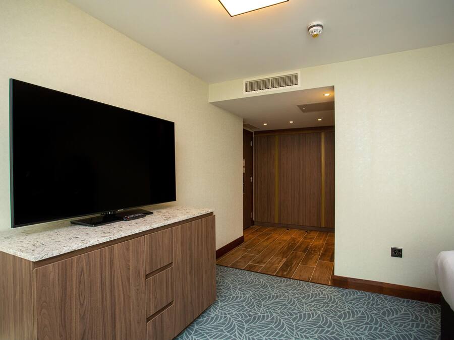 Habitación de hotel con televisor de pantalla plana sobre un mueble de madera, alfombra estampada y una cama.