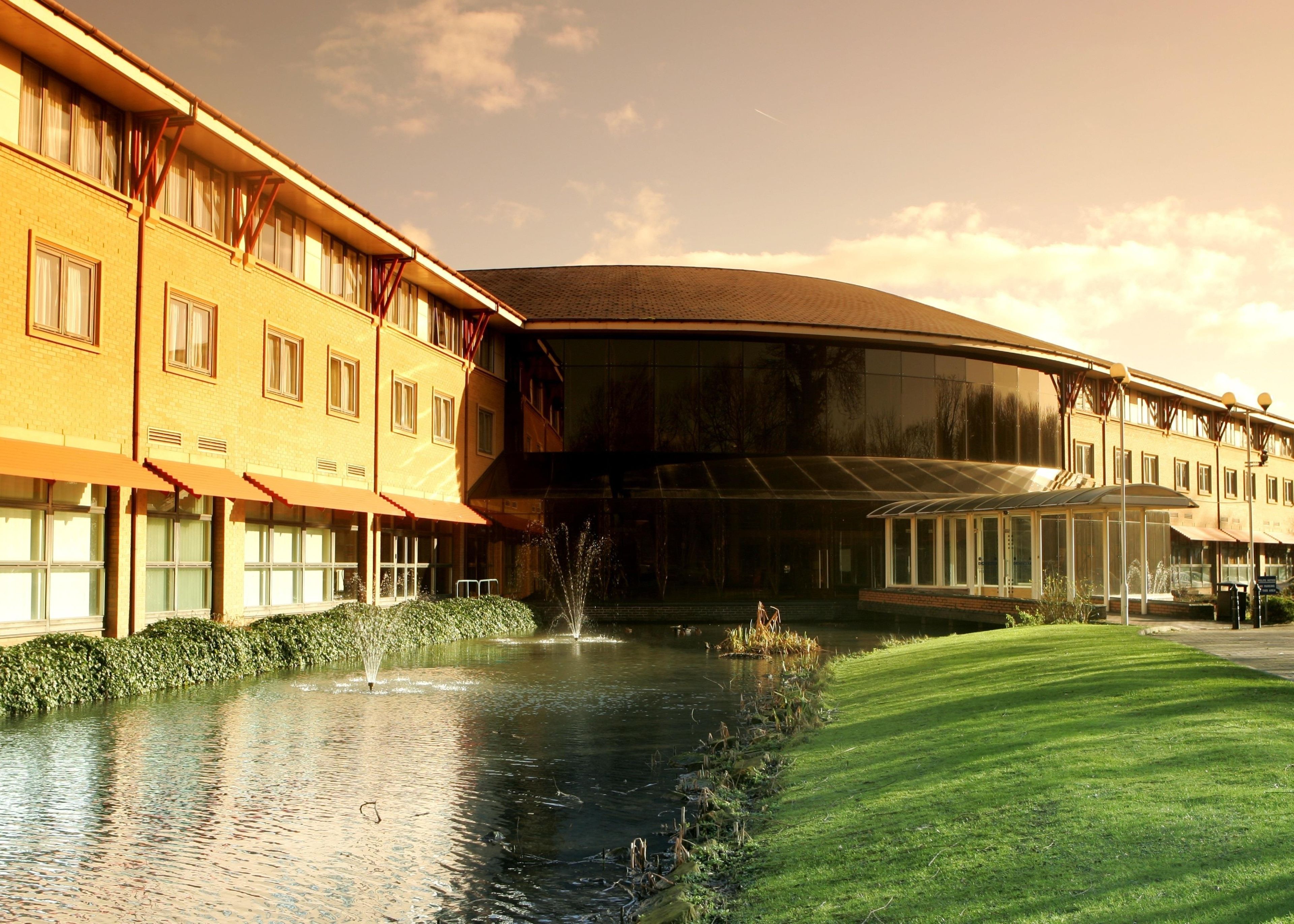 A hotel with a reflective glass facade, a landscaped lawn, and a water feature.