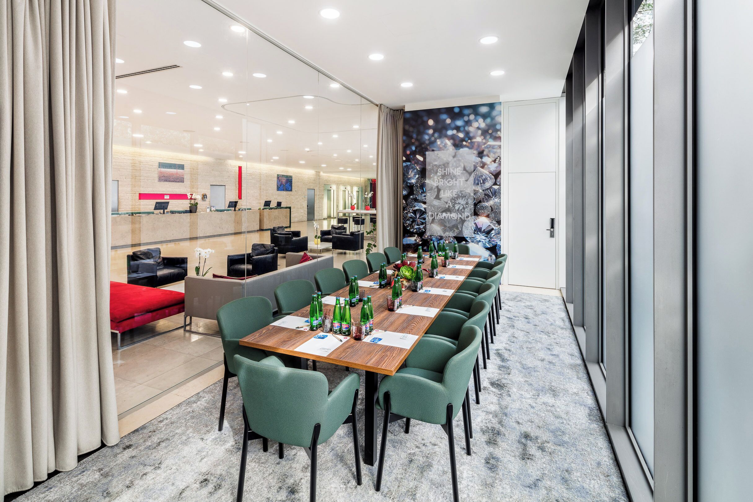 Un lobby moderno de hotel con una larga mesa de conferencias, sillas verdes y una zona de recepción al fondo.