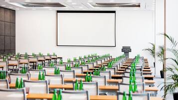 Un salón de conferencias con filas de mesas y sillas, una pantalla grande y un cartel que indica el nombre de la sala.