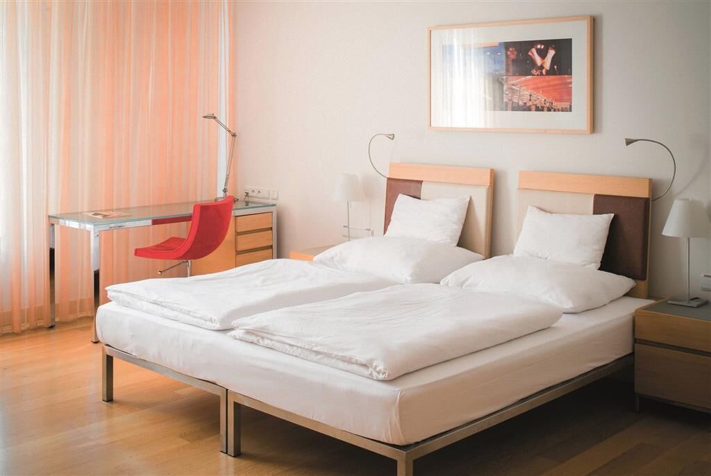 Una cama doble con sábanas blancas, un escritorio con una silla roja y una mesita de noche con lámpara.