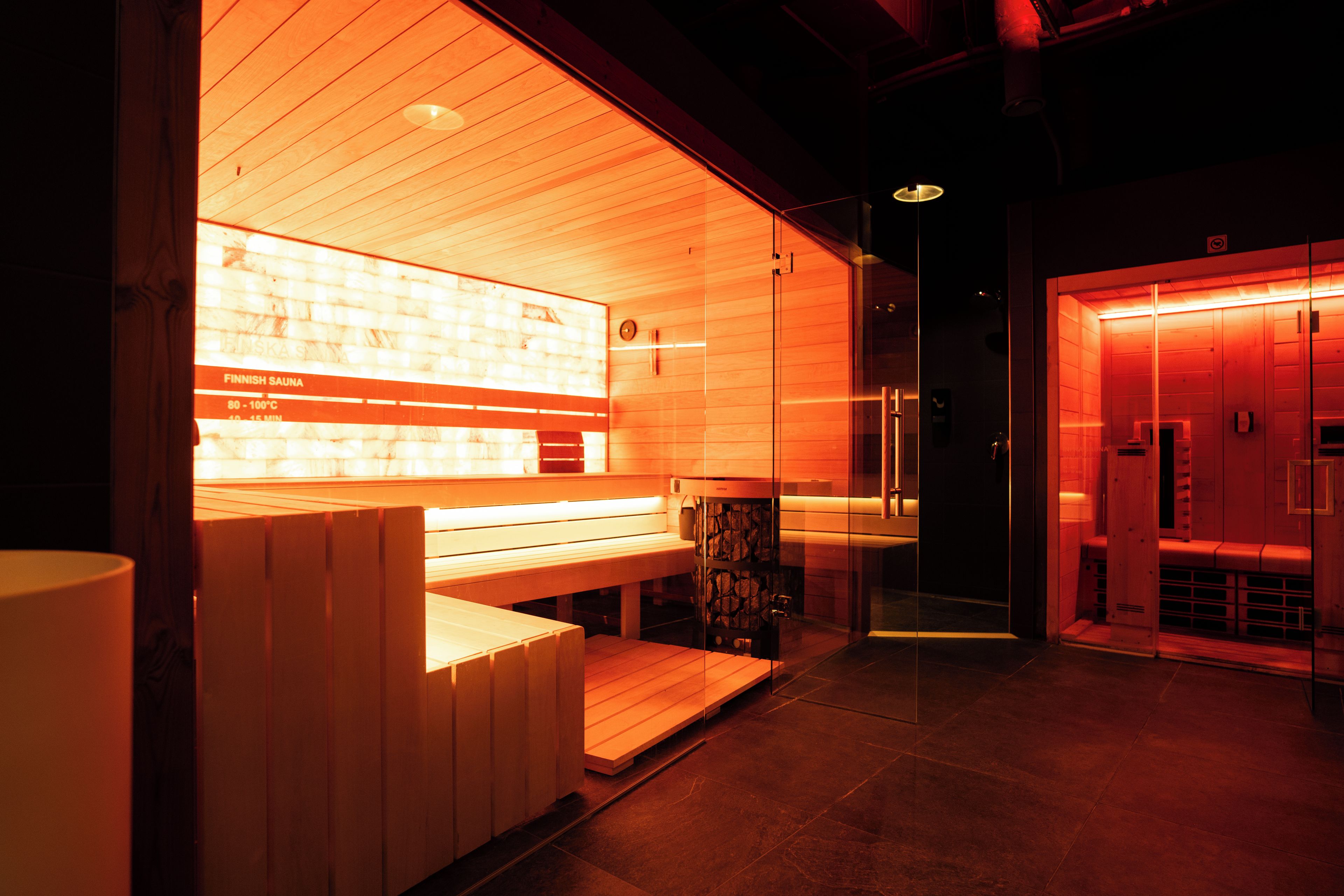 Uma sauna com bancos de madeira e divisória de vidro, painel de controle de temperatura visível e parede iluminada em vermelho.
