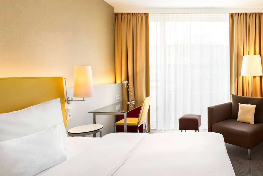 Quarto de hotel com uma cama grande, uma escrivaninha com cadeira, uma mesa pequena, um sofá e uma janela com cortinas.