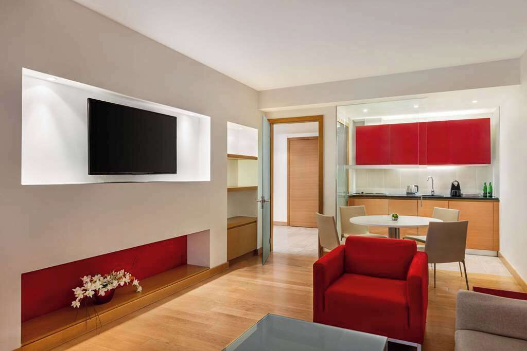 A modern living room with a flat-screen TV, a red sofa, a dining table, and a kitchen area with red cabinets.