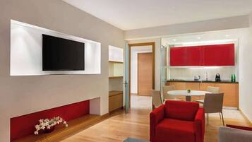 A modern living room with a flat-screen TV, a red sofa, a dining table, and a kitchen area with red cabinets.